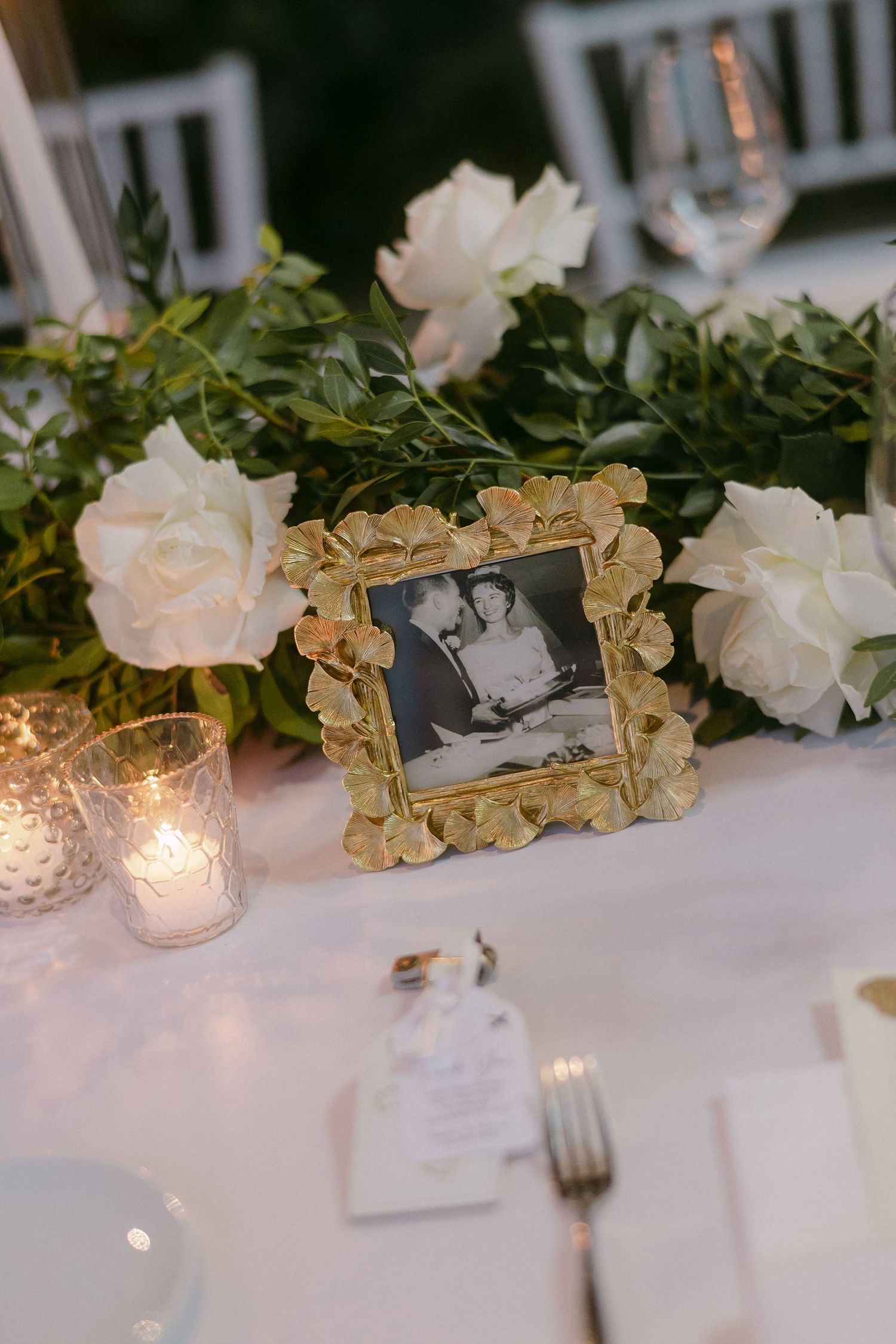 Elegant wedding table decor with ornate gold frames, white roses, and glowing candlelight on a white tablecloth.