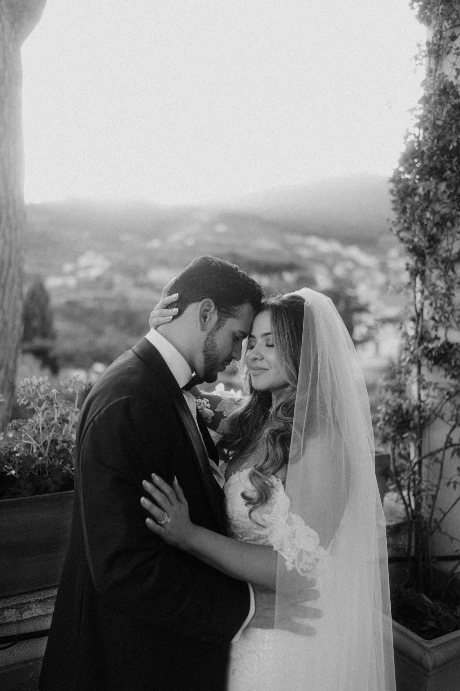A romantic black and white wedding photo showing a tender embrace between newlyweds on a hilltop overlook.