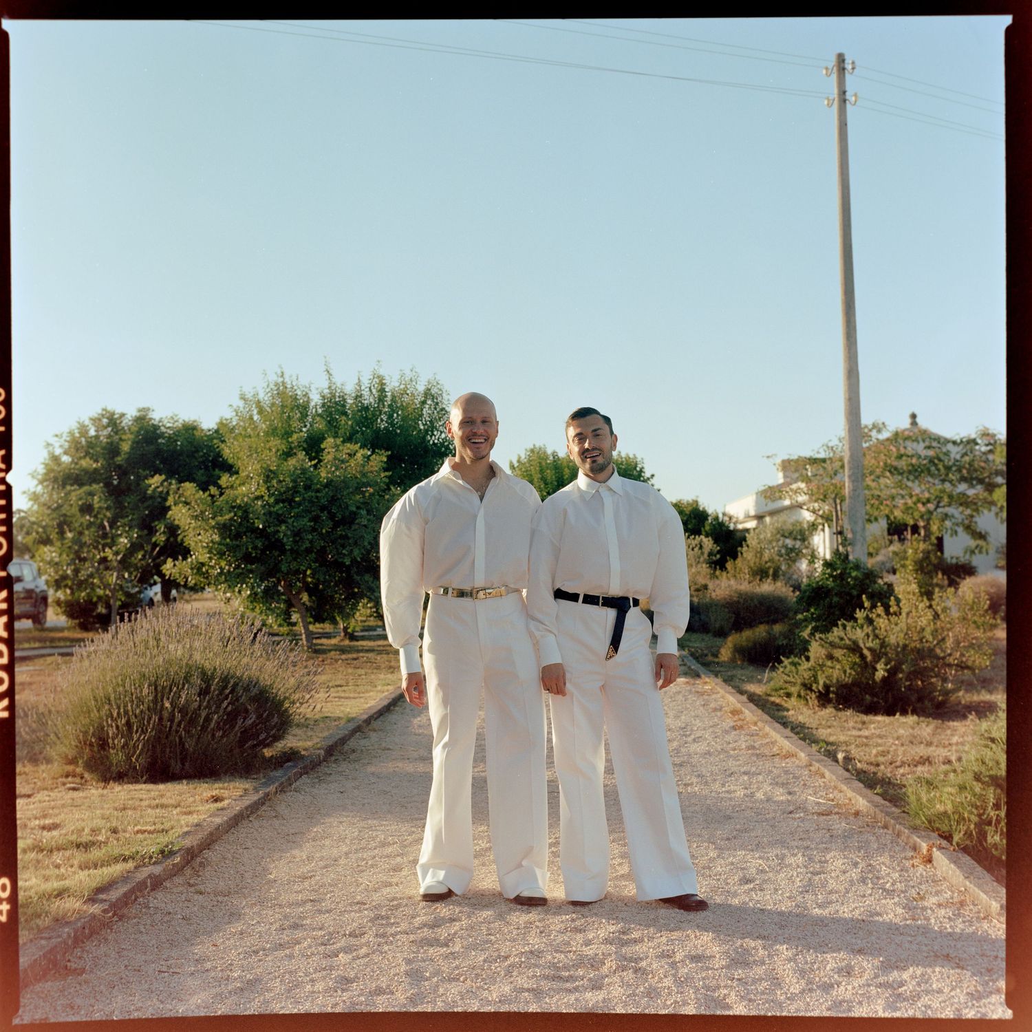 Two people stand on a dirt path wearing matching white outfits against a sky and desert landscape background.
