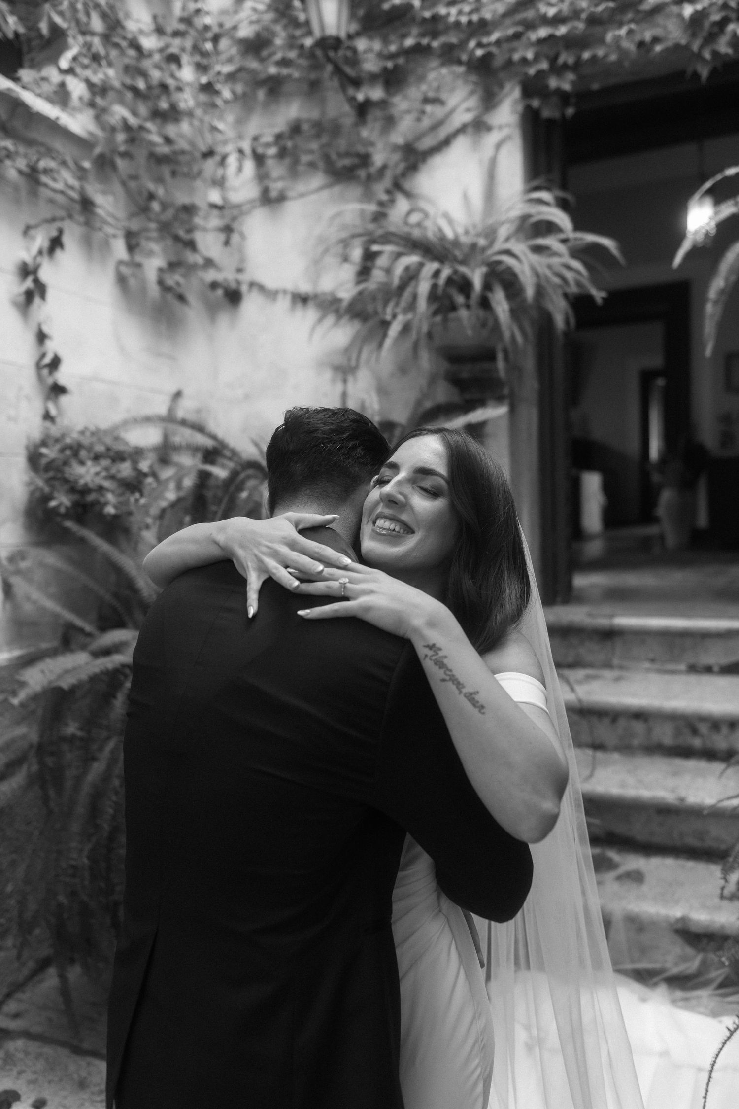 Black and white wedding photo showing a joyful embrace between newlyweds in a garden courtyard setting.