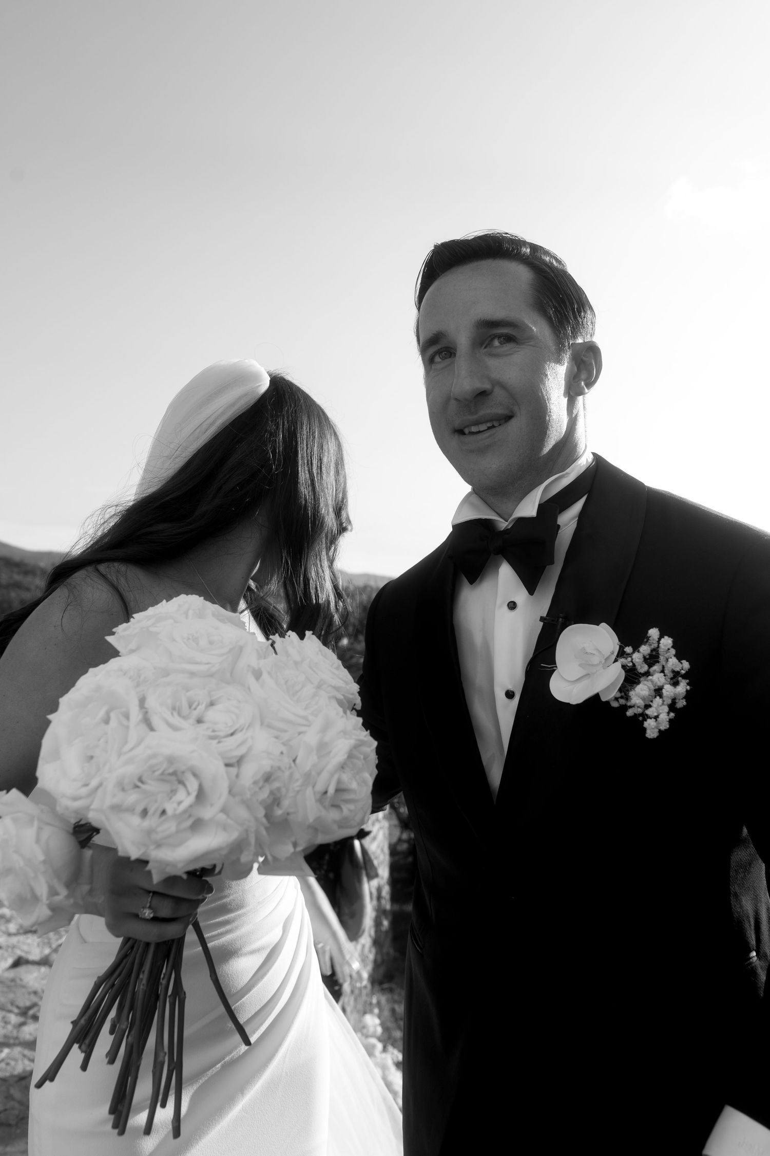A black and white wedding photo showing a bride holding white roses and groom in tuxedo during their ceremony.