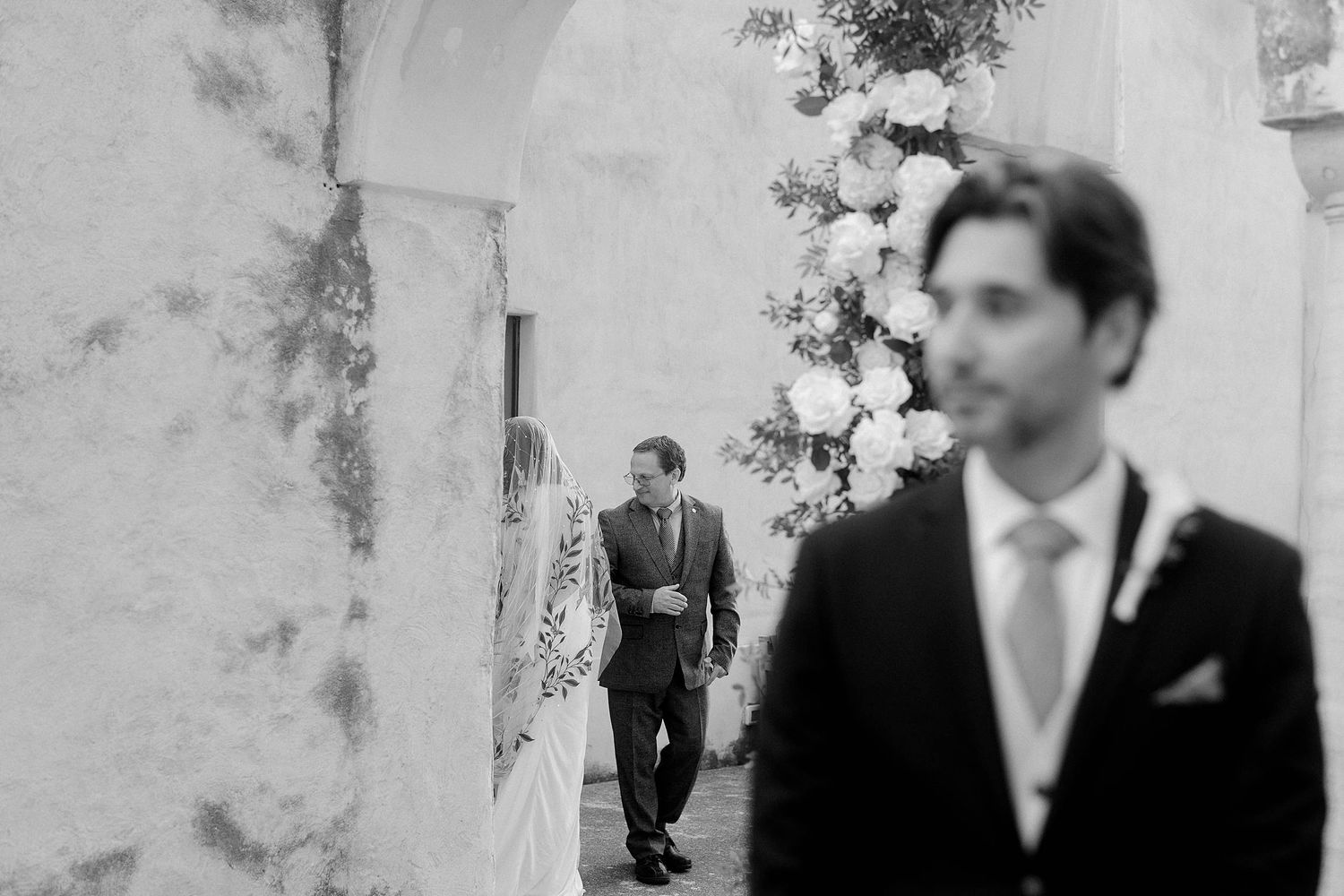 Black and white wedding photo showing a suited figure in foreground while another walks through an arched doorway.