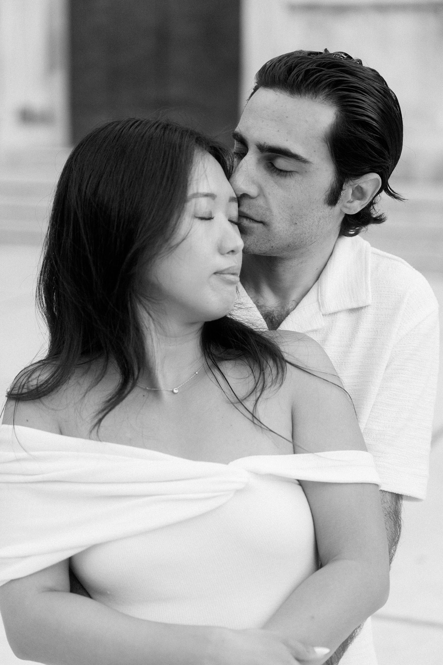 Black and white romantic portrait of an intimate couple in white clothing embracing tenderly against a light background.