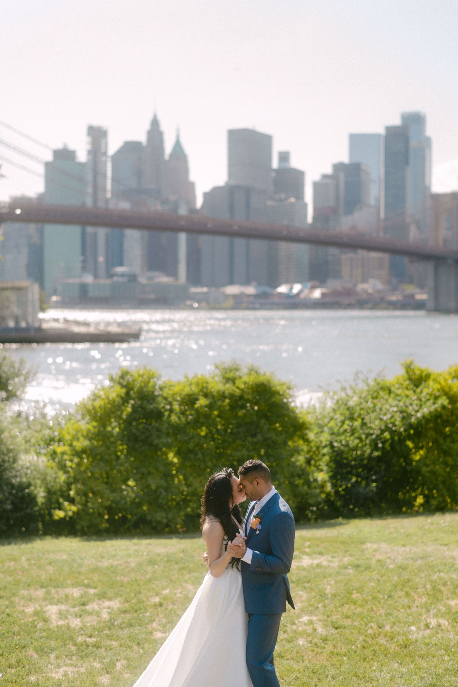 A romantic wedding couple shares a tender moment on a grassy hill overlooking the Manhattan skyline on a sunny day.