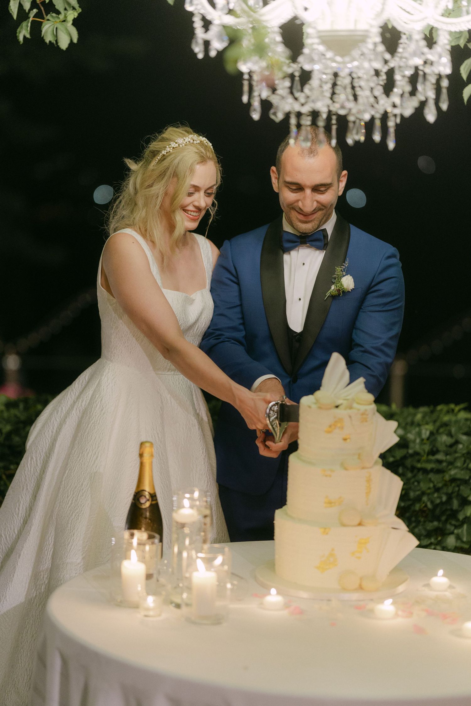 A couple in formal wedding attire cuts their elegant white wedding cake surrounded by glowing candles at an evening reception.