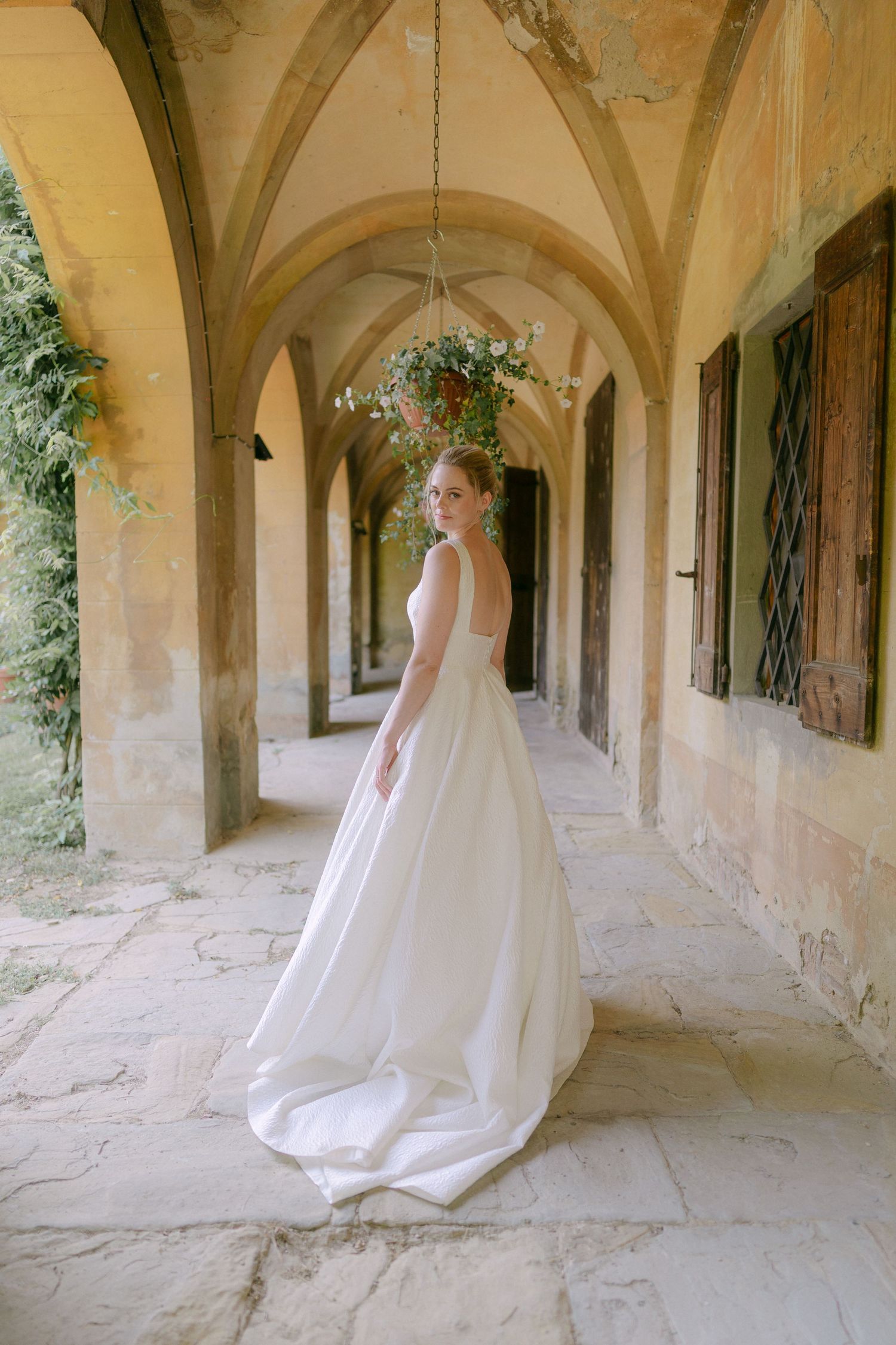 A flowing white wedding dress sweeps across a stone colonnade hallway in an elegant architectural setting.