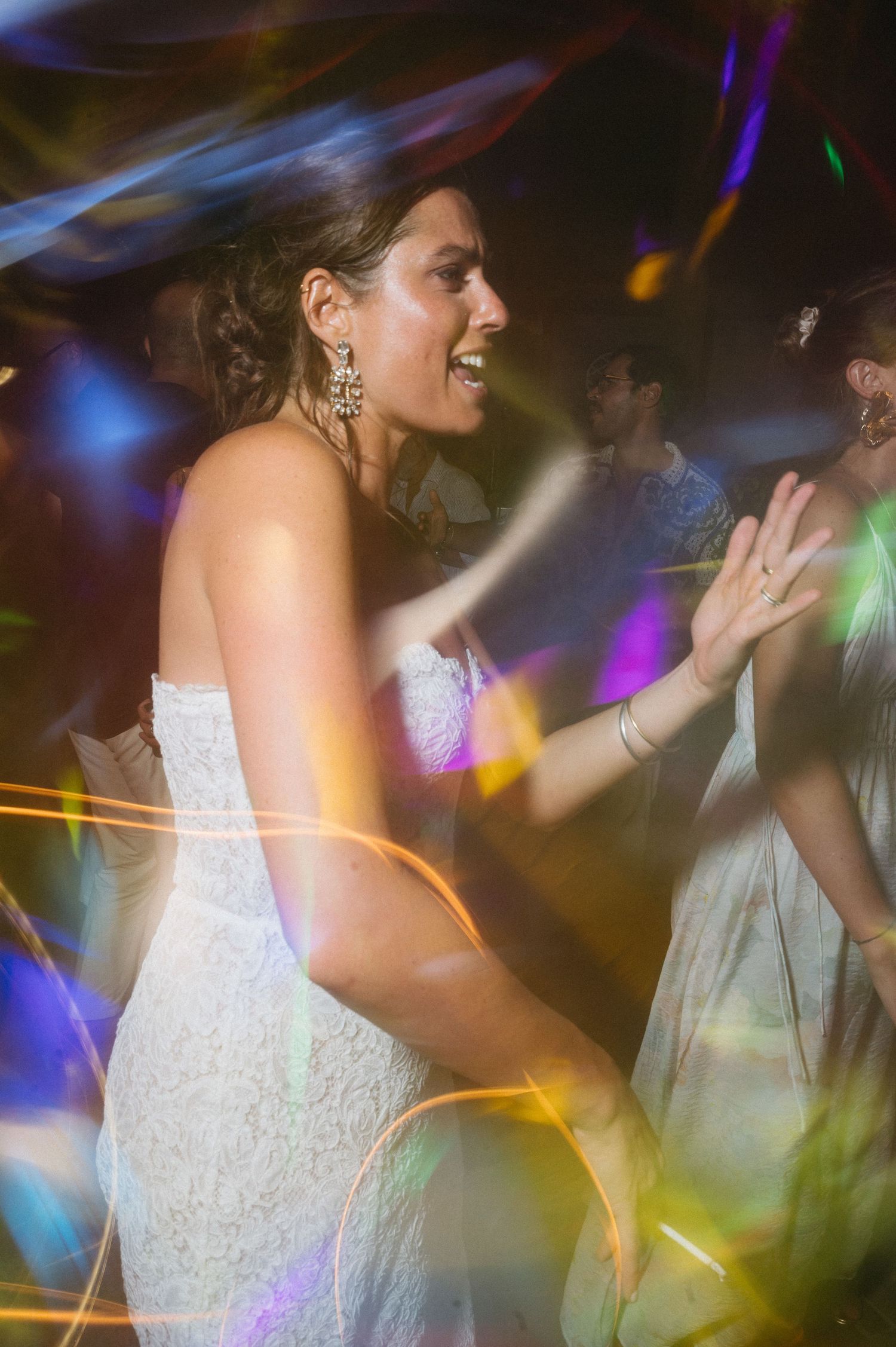 A bride in a white lace dress dances joyfully at a wedding reception with colorful lights swirling around her.