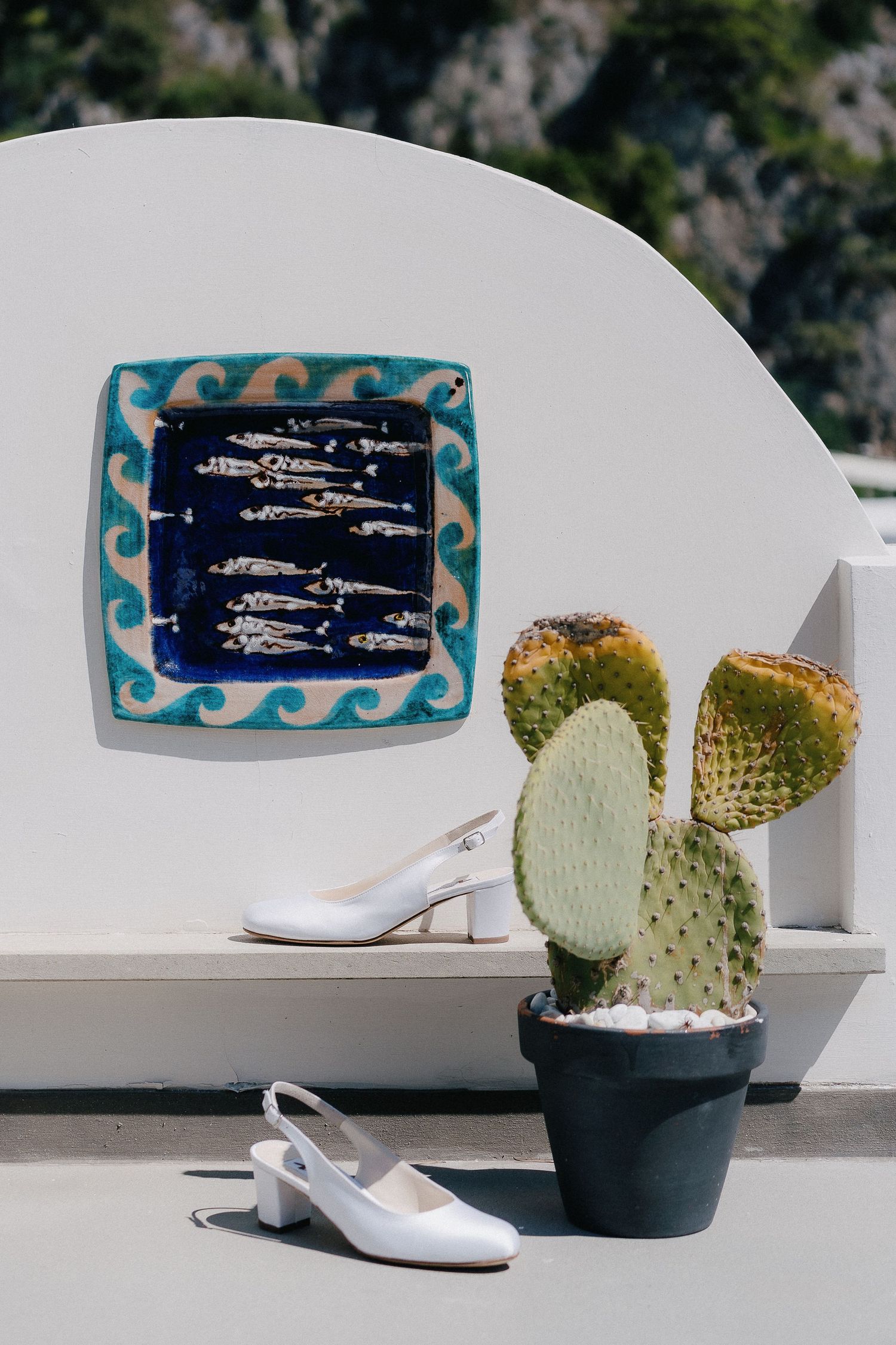 White slingback shoes placed near a potted cactus plant and decorative fish artwork on a Mediterranean terrace.