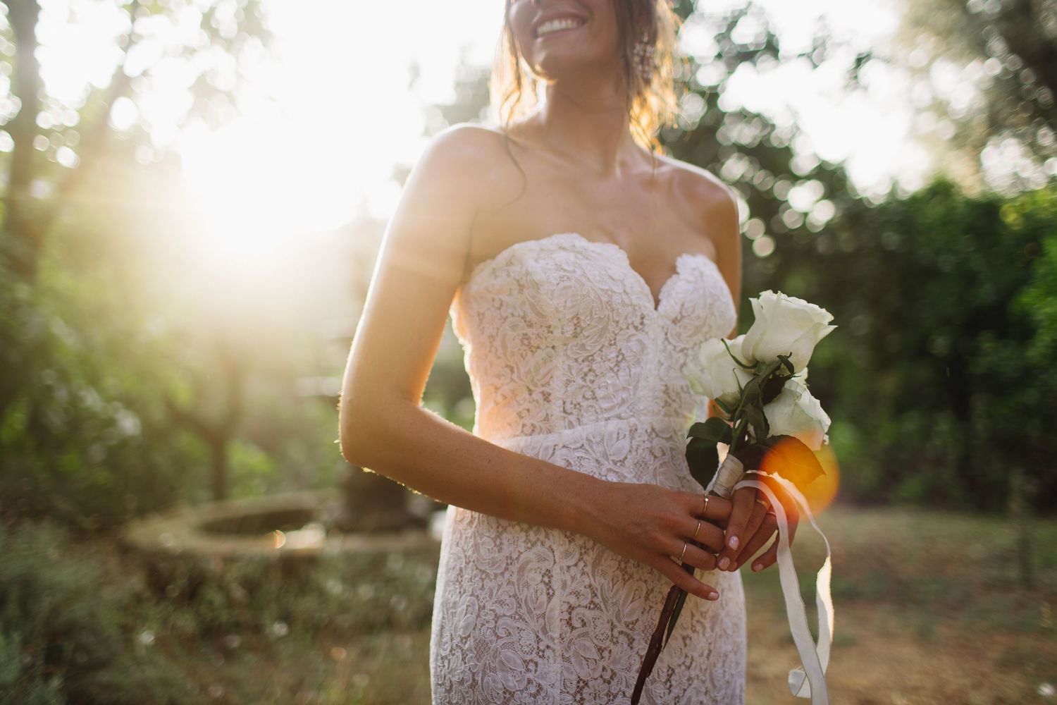 Bride holding a white floral bouquet in a lace wedding dress during golden hour sunset outdoors.