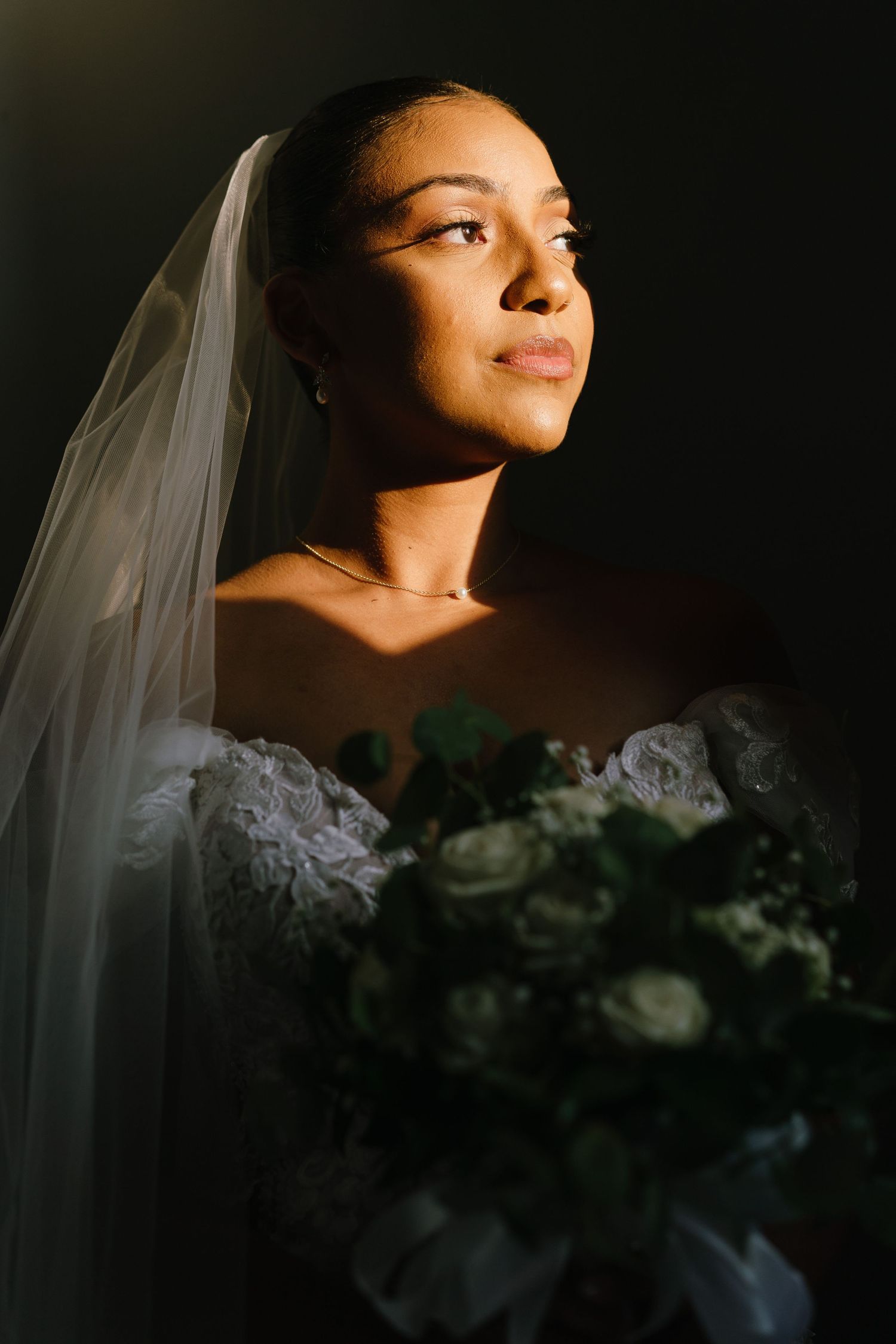 A bride in dramatic side lighting poses with white roses and veil creating an elegant bridal portrait series.