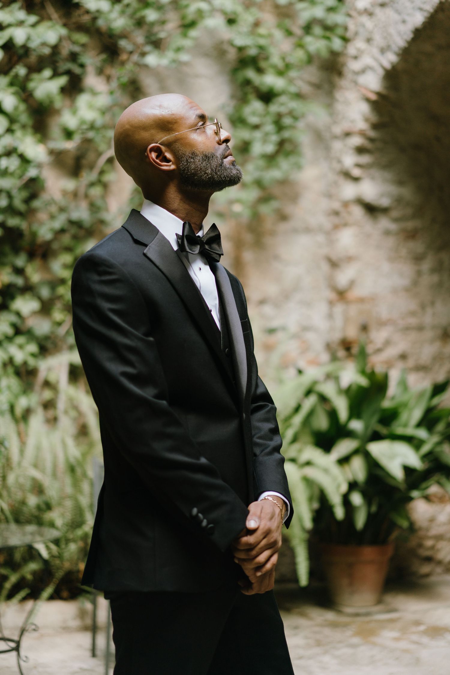 A person in a black suit and tie stands thoughtfully against a garden wall with potted plants in the background.