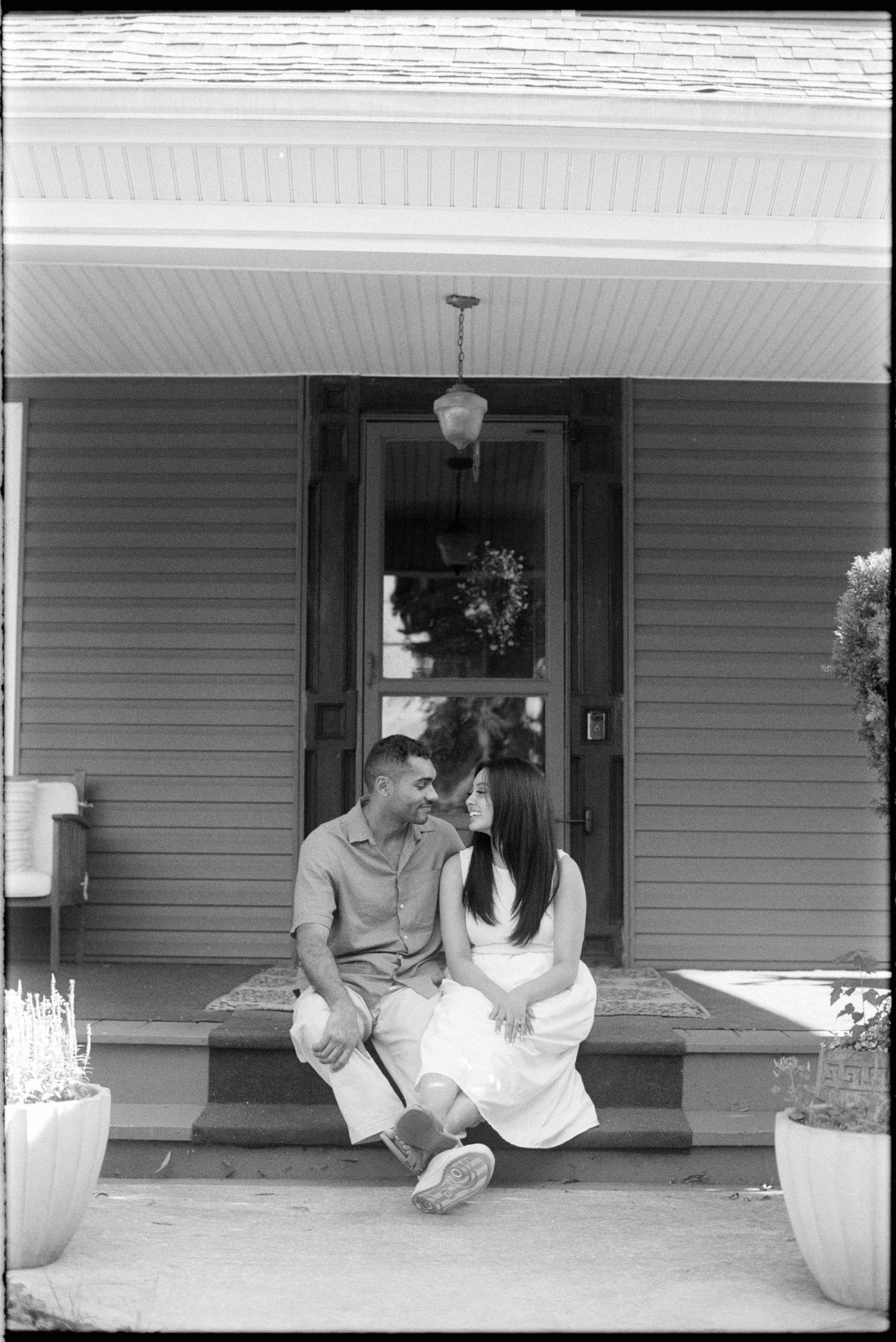 A couple sits together on a front porch step beneath a hanging light fixture and decorative wreath on the door.