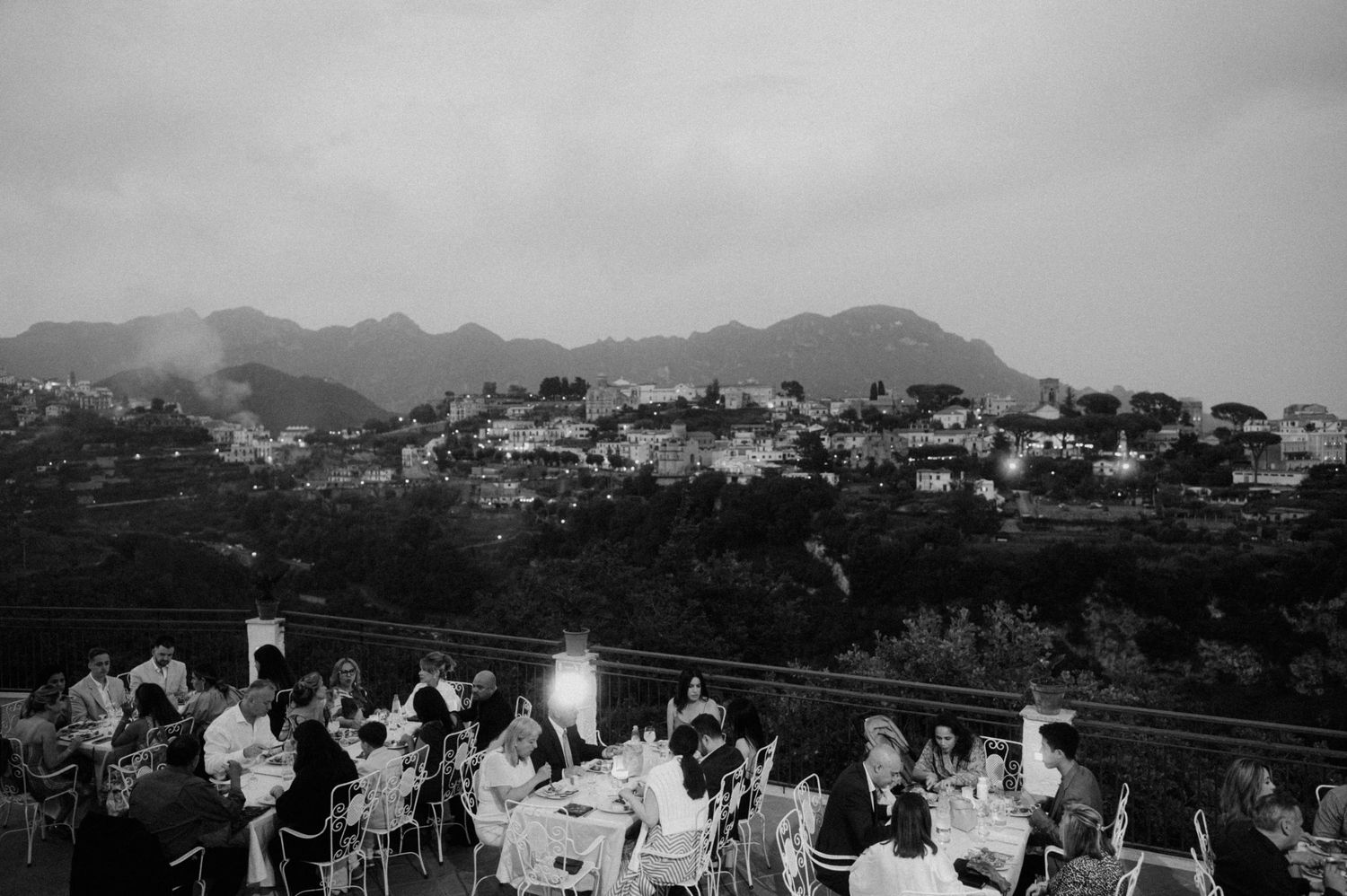 People gather at an outdoor terrace overlooking mountainous coastal town in black and white photography.