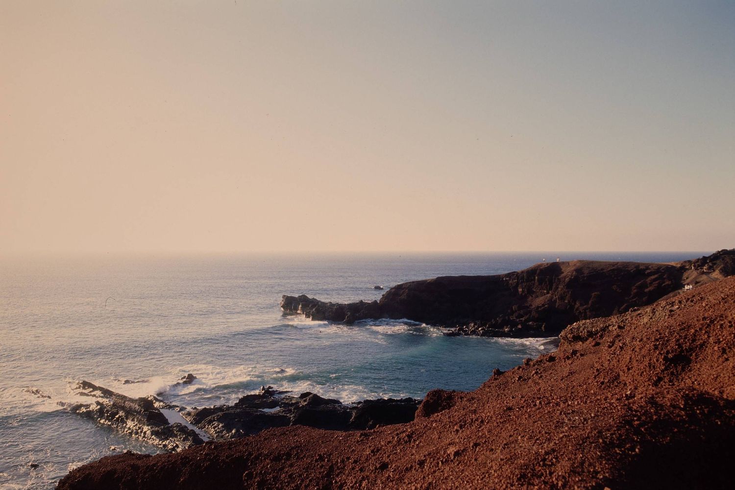 Dramatic coastal cliffs and rugged shoreline at sunset overlooking a turquoise ocean bay under a hazy sky.