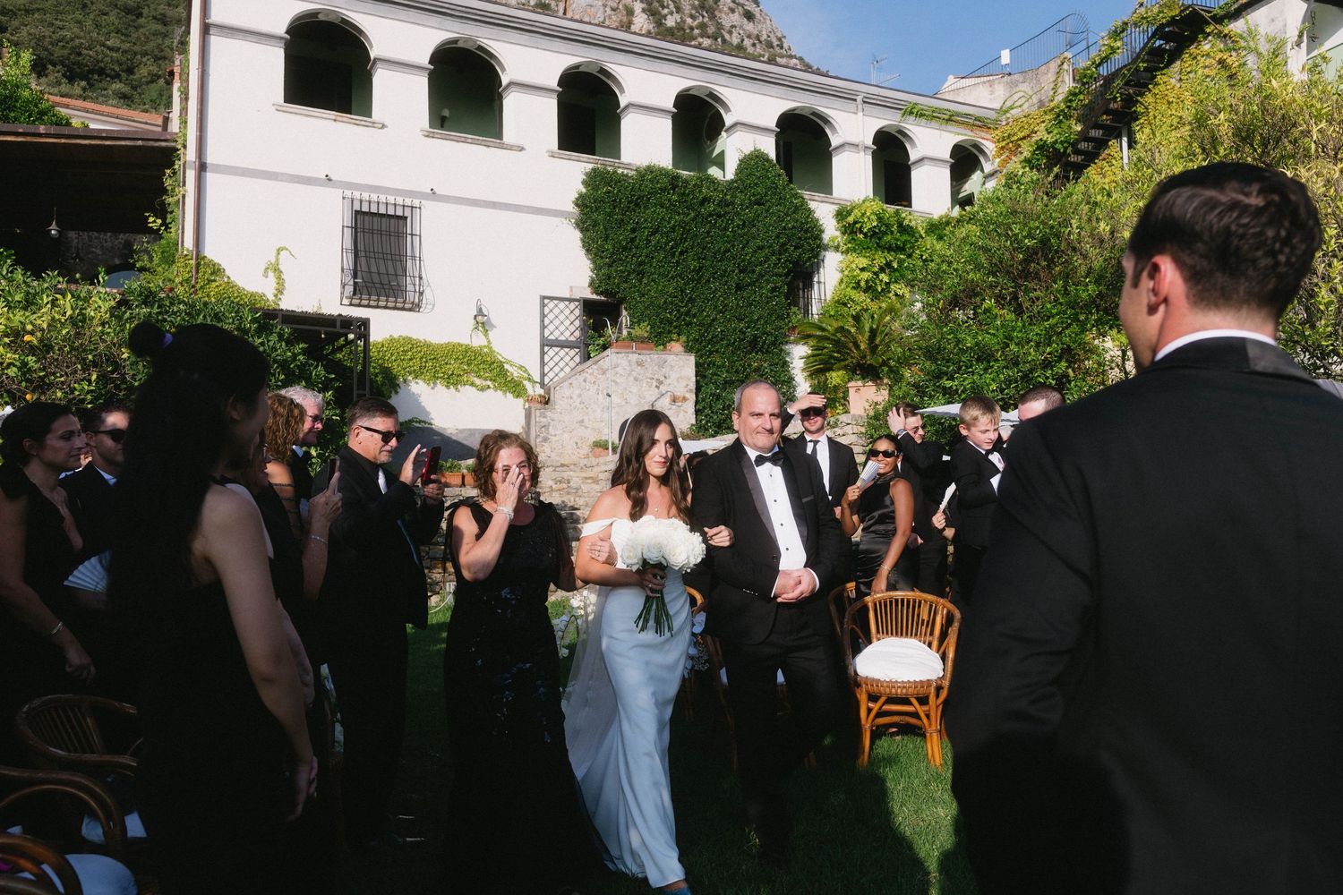 Wedding guests gather outdoors in a garden courtyard surrounded by ivy-covered walls during a celebration.