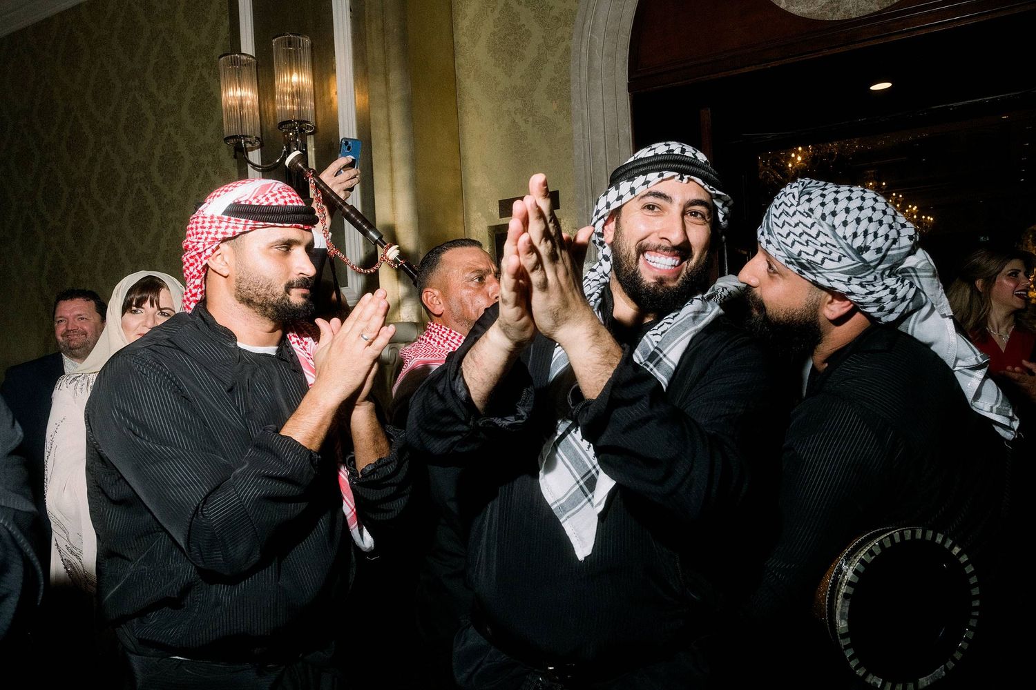 Celebratory group of men wearing black and white traditional Middle Eastern headscarves clap together at formal event.