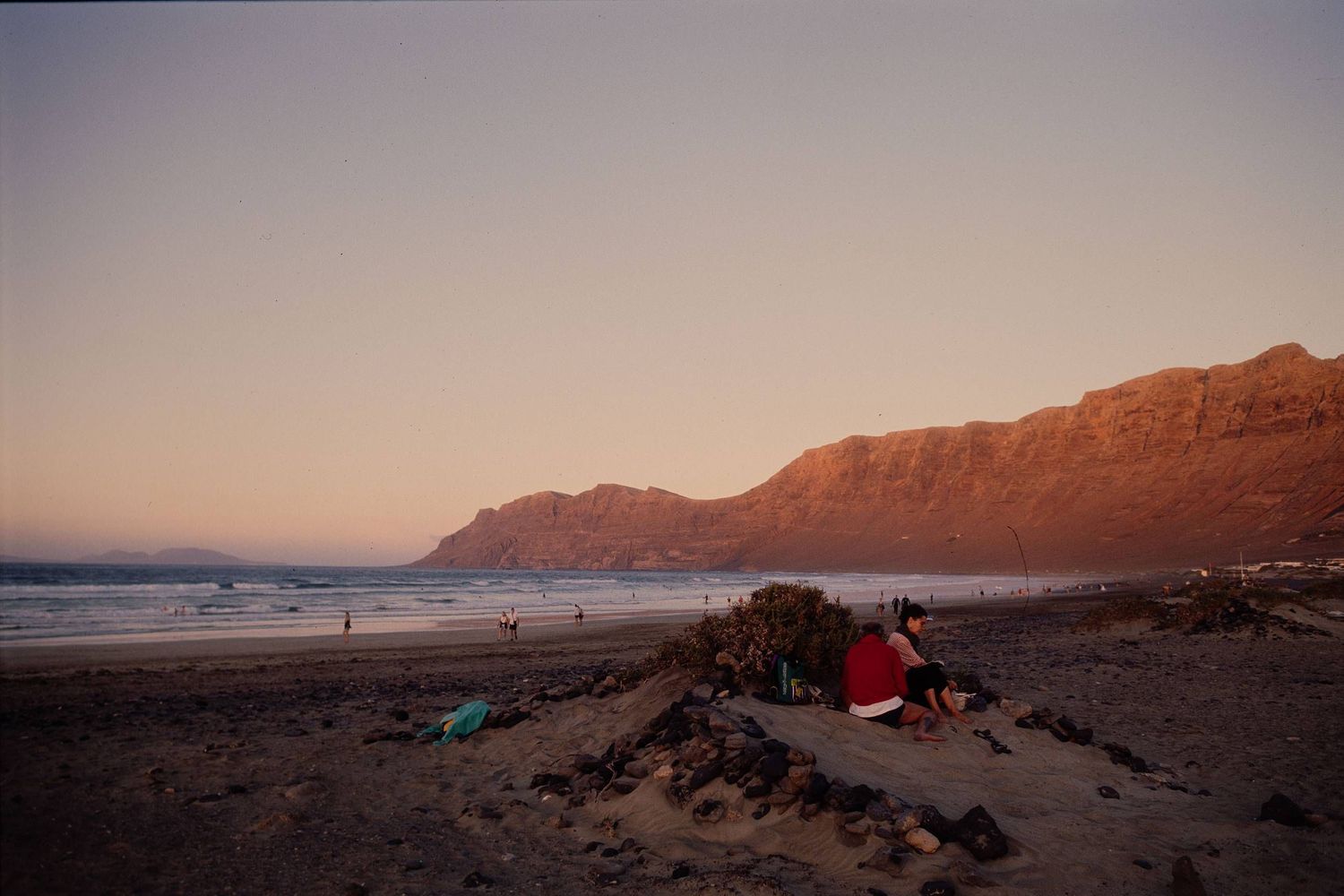Red tent on sandy beach at sunset with mountain cliffs and ocean waves in background.