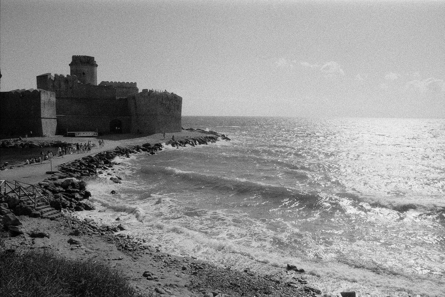 A black and white coastal landscape shows waves crashing against rocky shores near a medieval fortress wall.