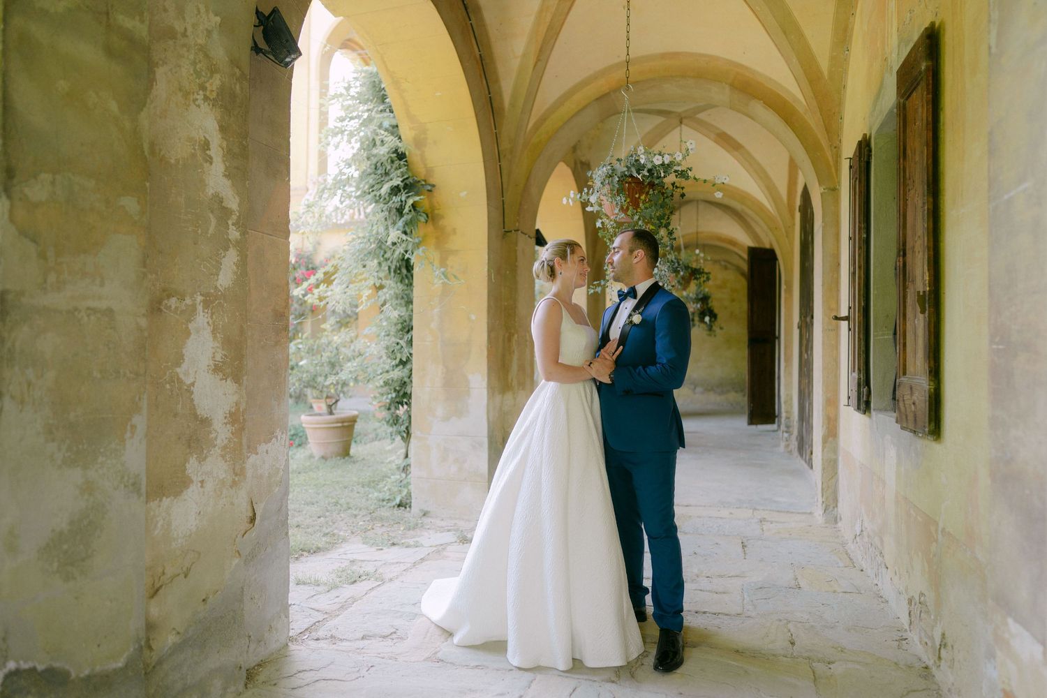 A romantic couple embraces in a stone archway corridor with natural lighting and architectural columns.