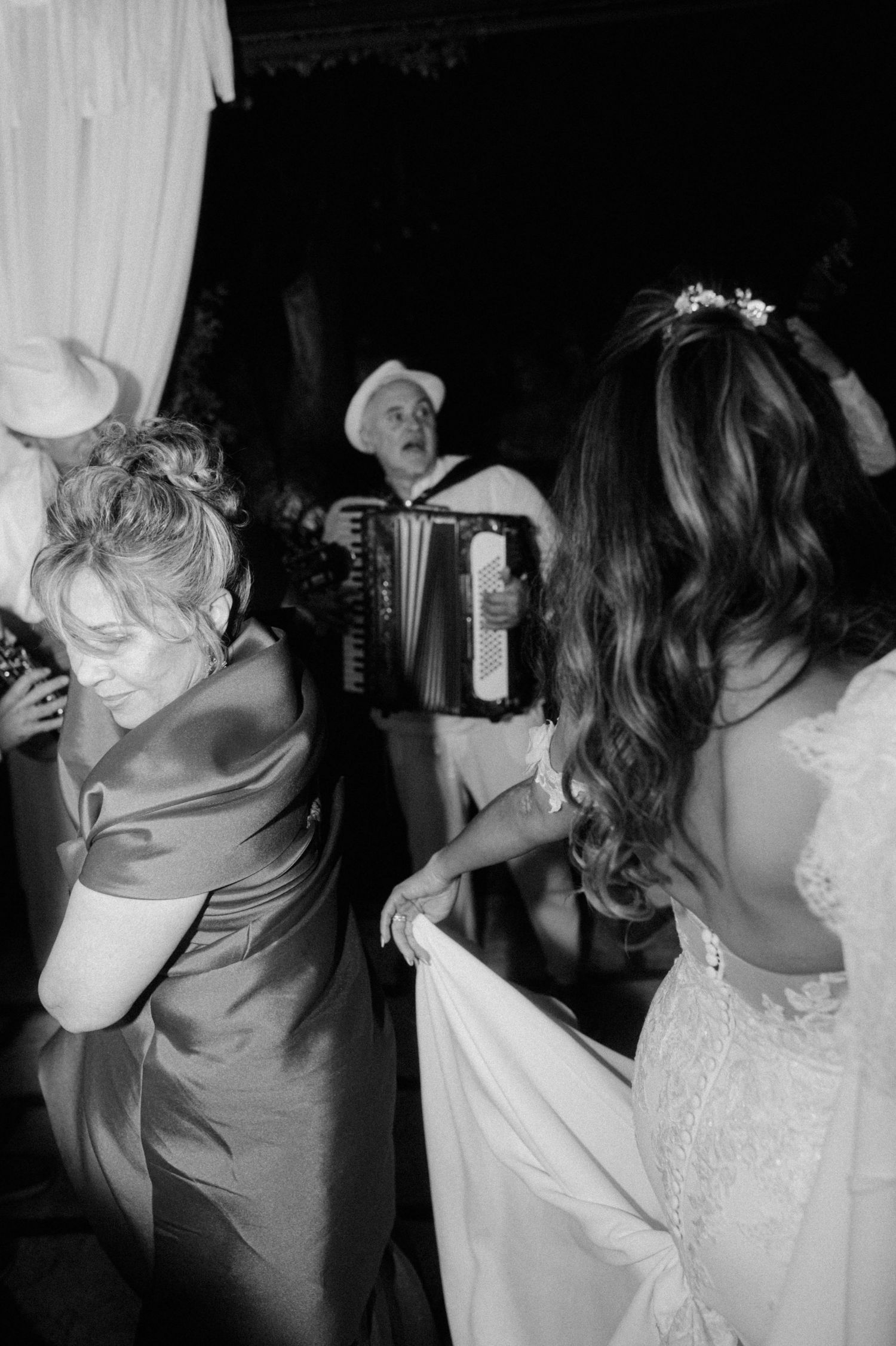 Black and white photo of people dancing at a wedding reception while an accordion player performs in the background.