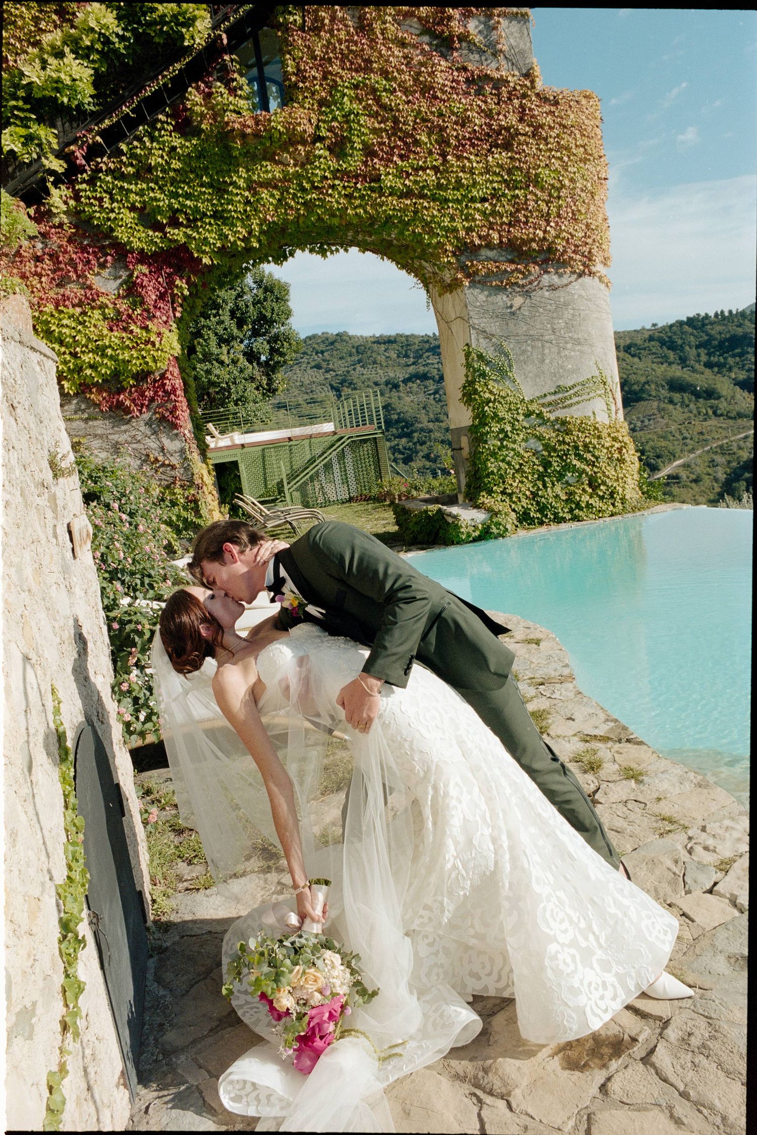 A romantic wedding photo at a scenic stone archway overlooking a turquoise pool with ivy-covered walls.