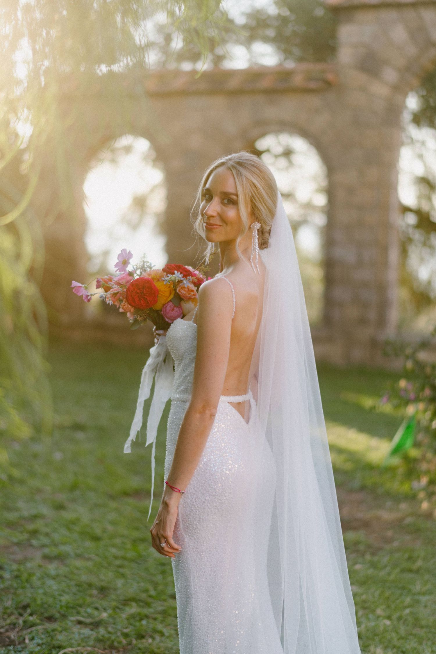 A bride in a backless white wedding dress holds a vibrant fall-colored bouquet in a sun-dappled garden setting.