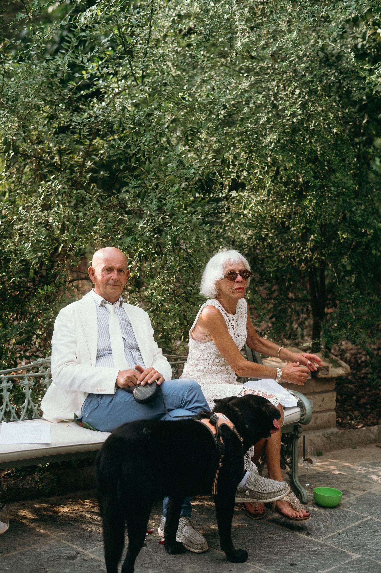 Two people sit together on a stone bench in a garden setting surrounded by lush greenery.
