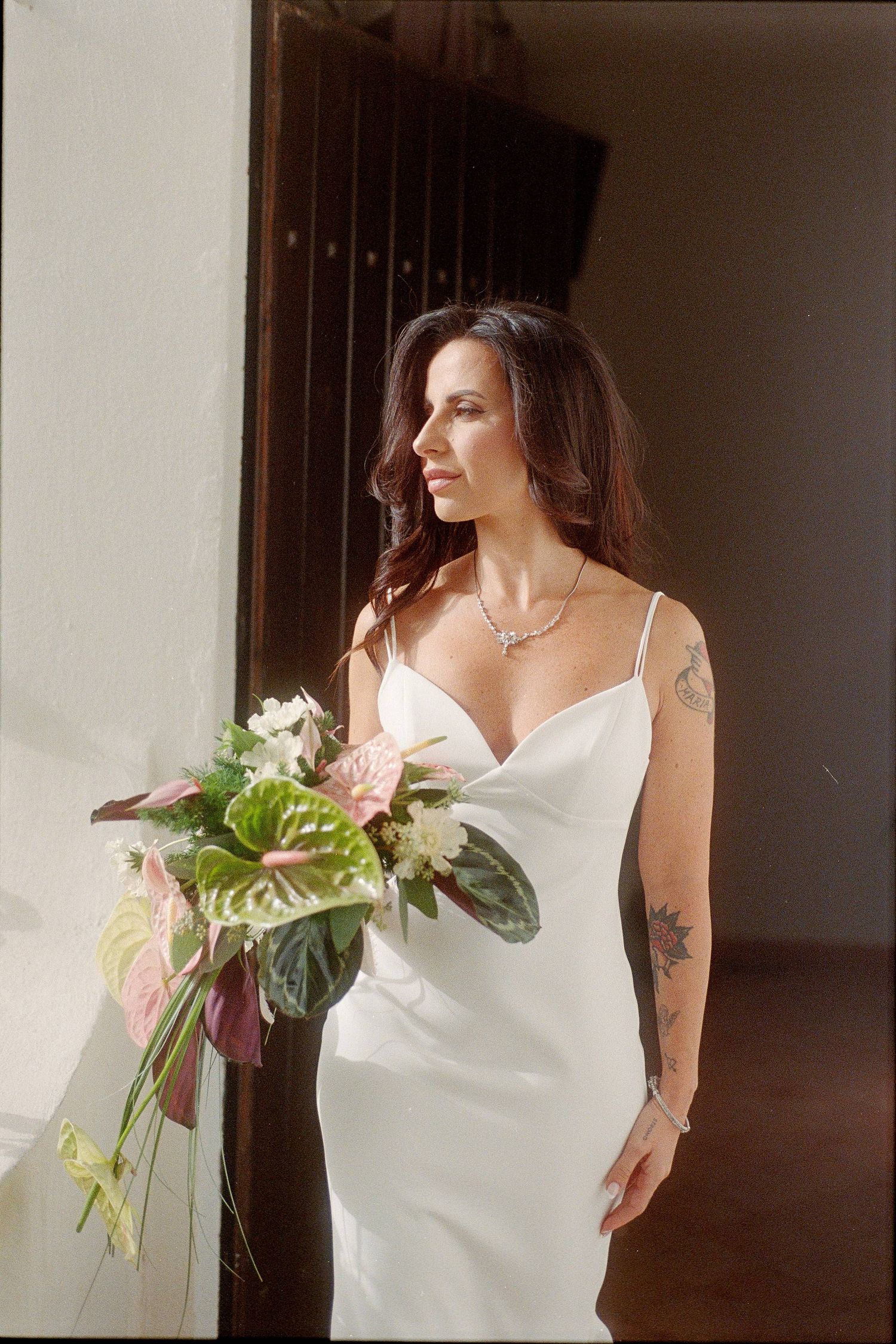 A bride in a white wedding dress holds a delicate bouquet of flowers while standing in natural window light.