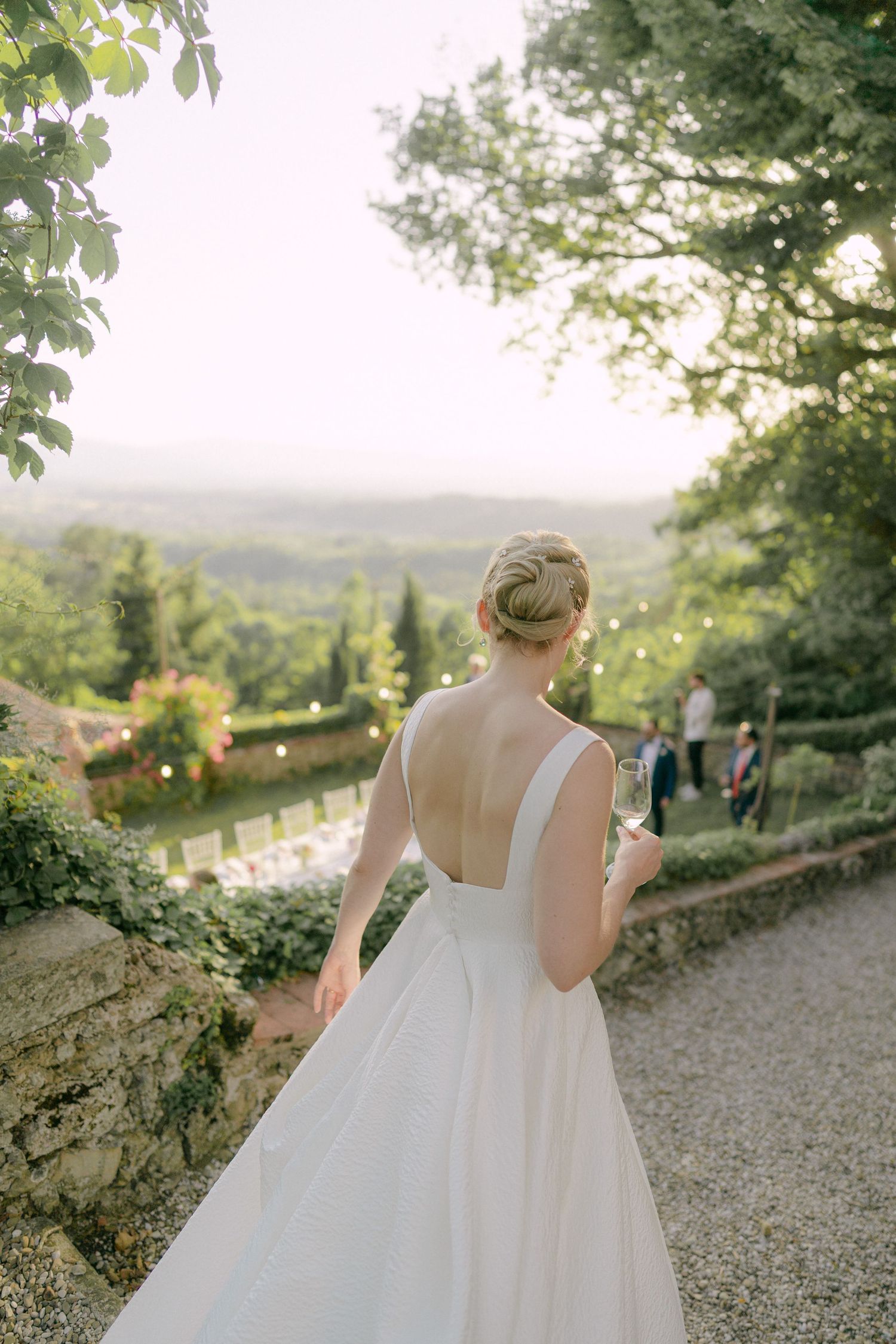 A bride in a flowing white dress stands on a gravel path overlooking a scenic Tuscan countryside at sunset.