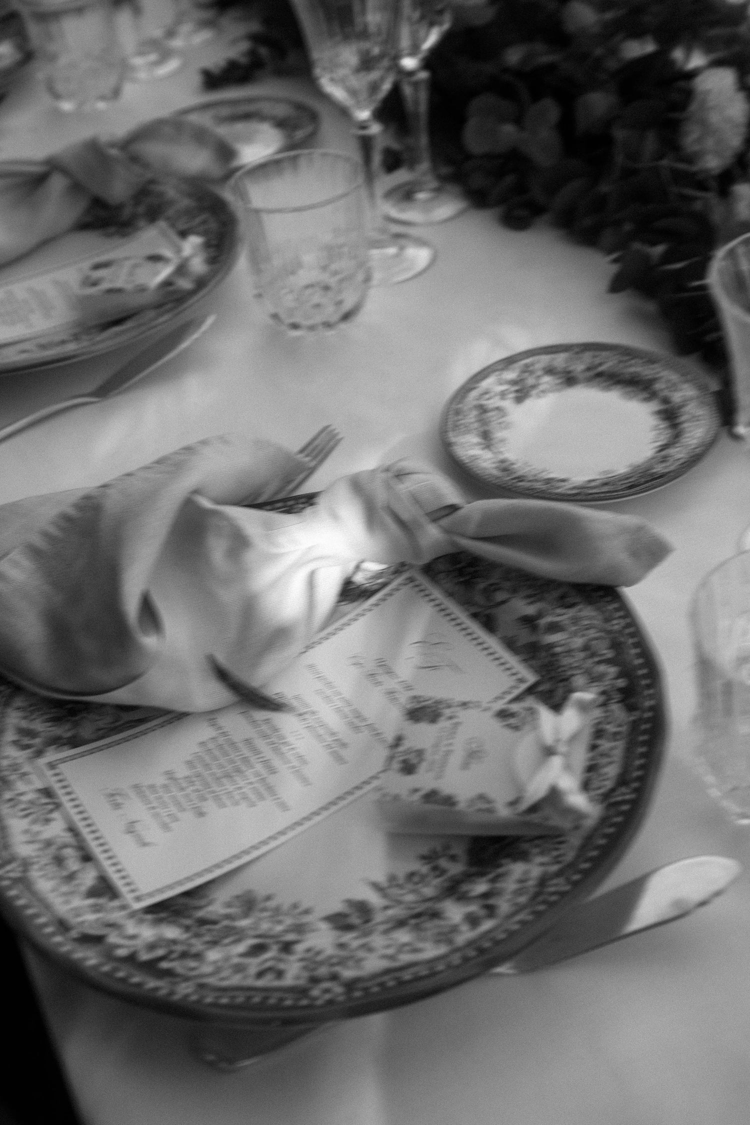 Black and white photo of an elegant table setting with ornate plates, folded napkin, menu card, and crystal glassware.