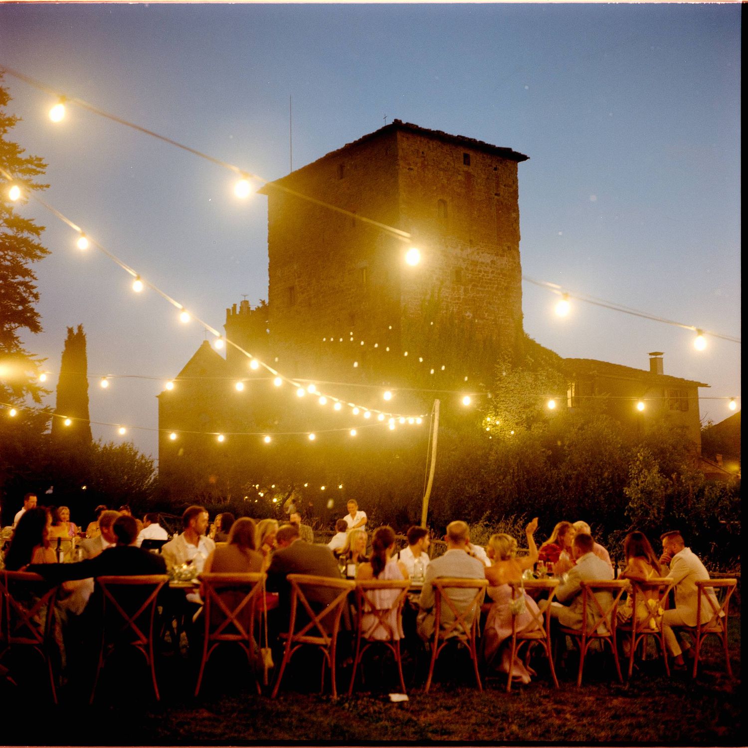 Outdoor evening dinner party under string lights near a historic stone castle tower at dusk.