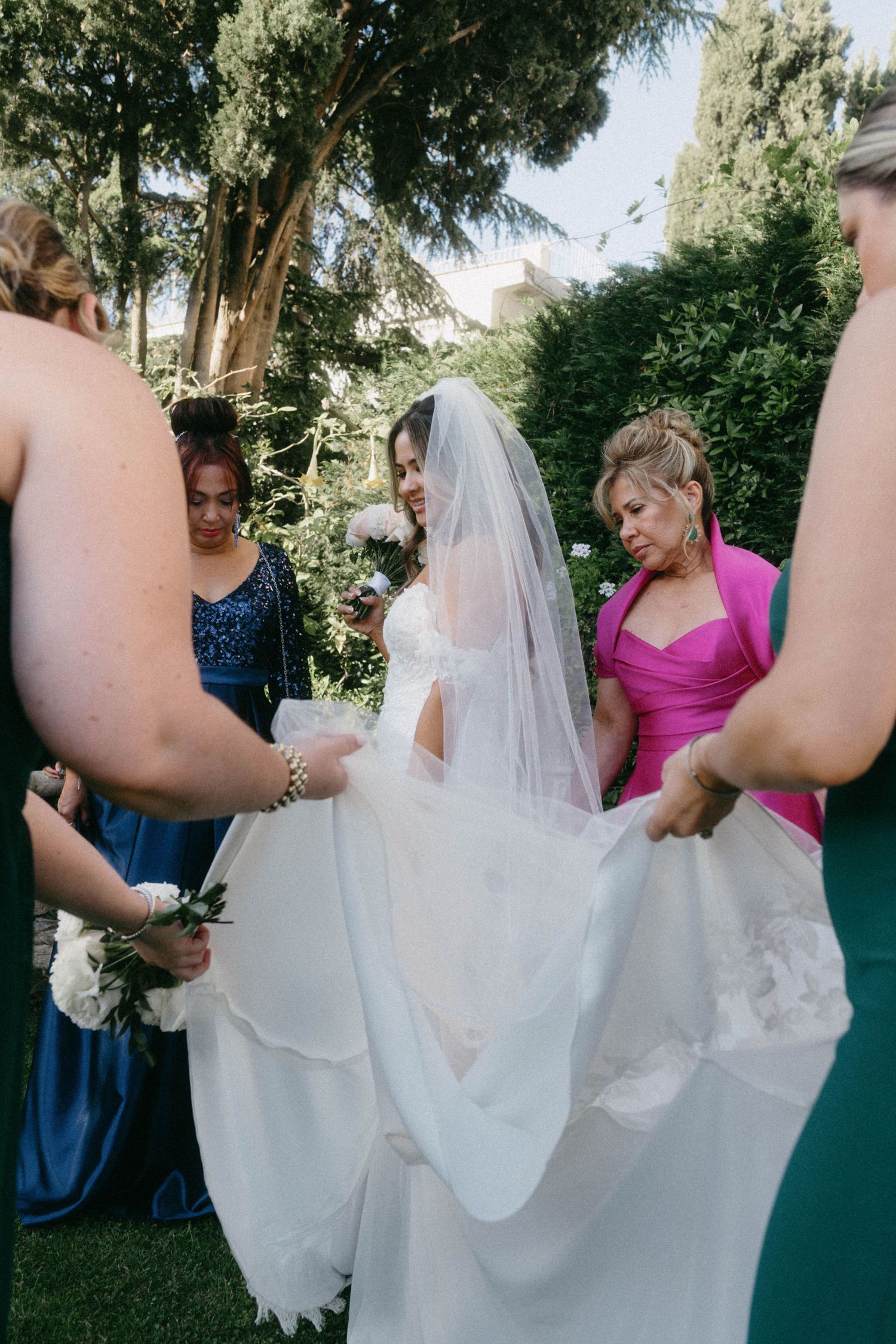 Bridesmaids in blue and pink dresses help adjust a bride's flowing white wedding gown and veil in an outdoor garden setting.