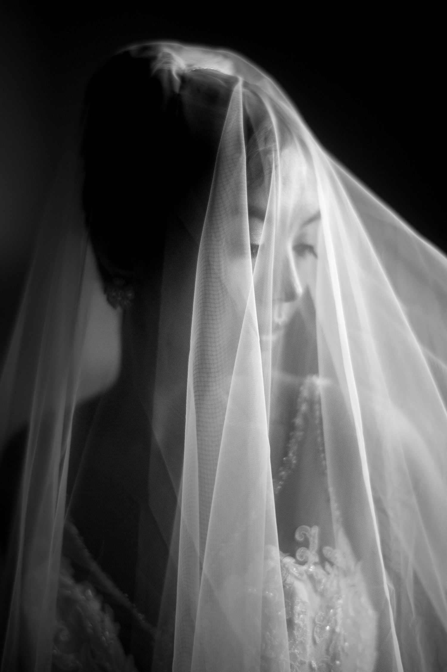 Artistic black and white wedding photo showing a figure beneath a delicate flowing veil creating a soft ethereal effect.