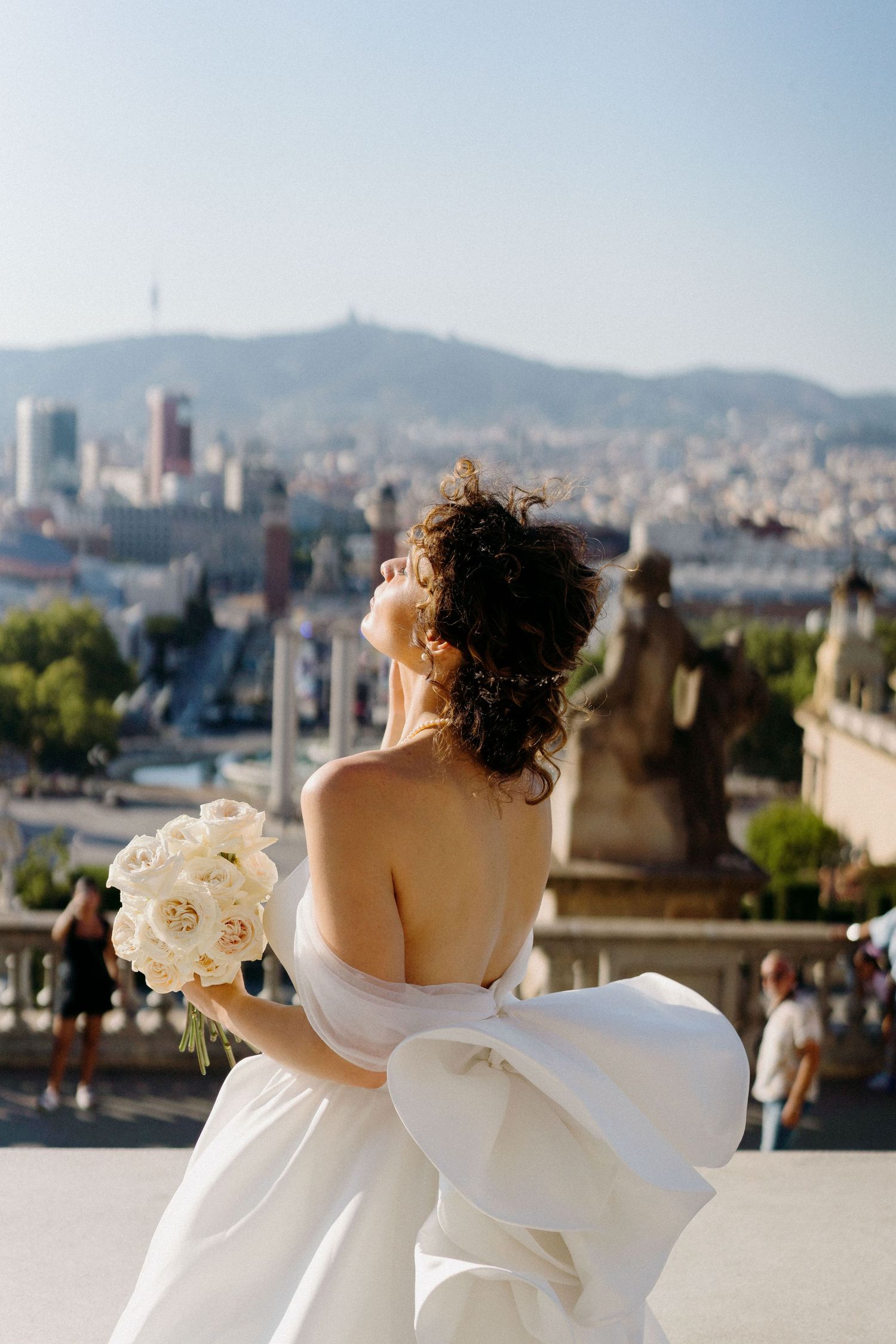 Bride in white dress holding roses looks over Barcelona cityscape from high viewpoint during golden hour.