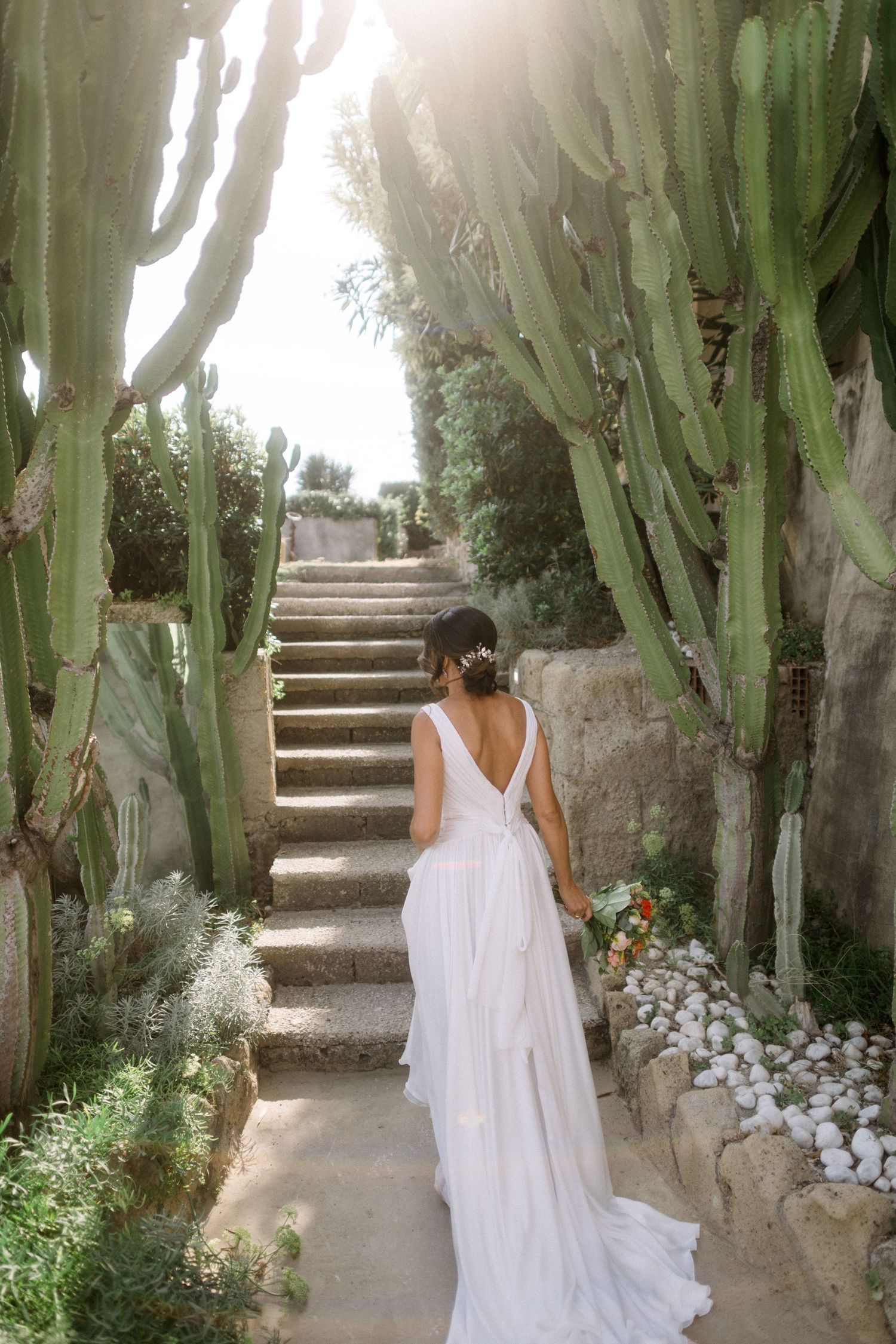 A flowing white wedding dress with a low back is seen on a stone stairway lined with tall cacti and florals.