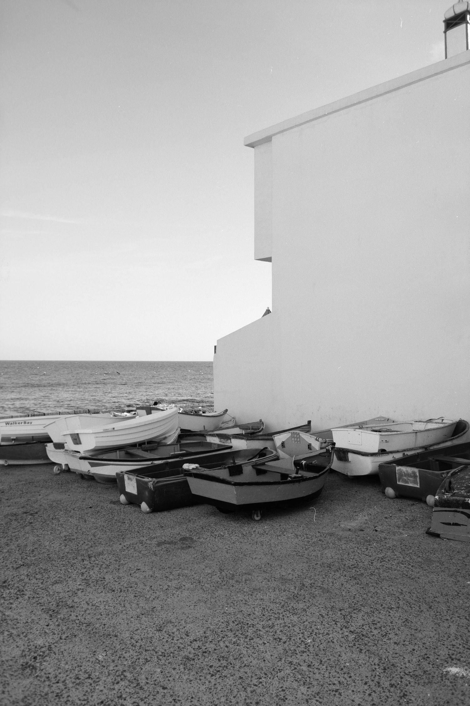 Small fishing boats lined up against a white building wall near the seashore in black and white photography.