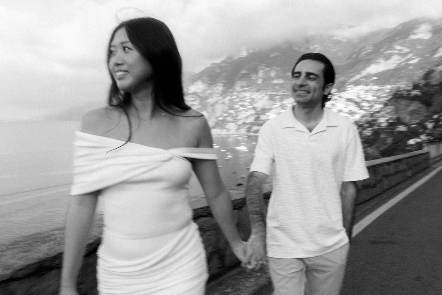 A couple in white outfits walks along a scenic coastal road with mountains in the background.