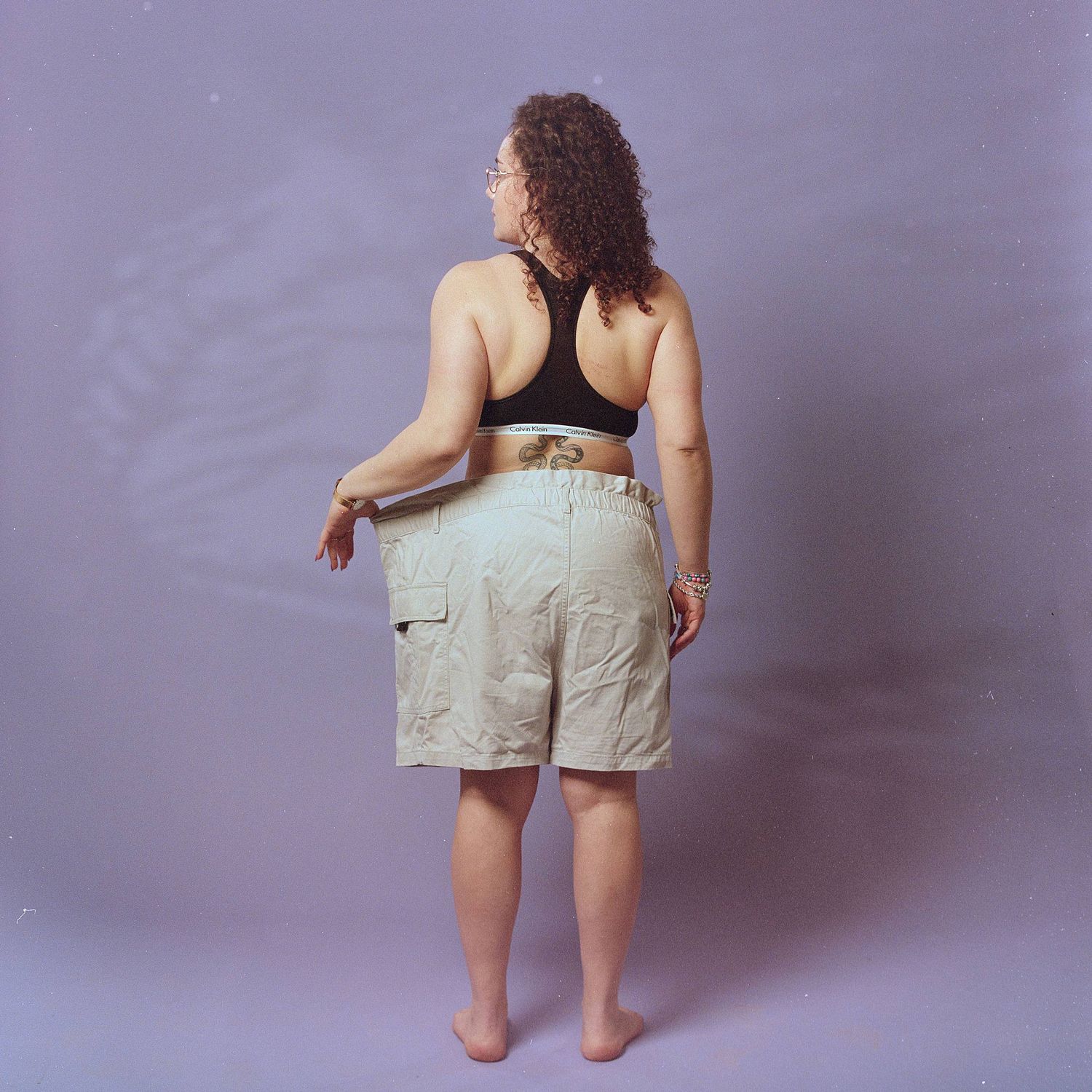Rear view of person wearing black sports bra and beige cargo shorts against purple background in studio setting.