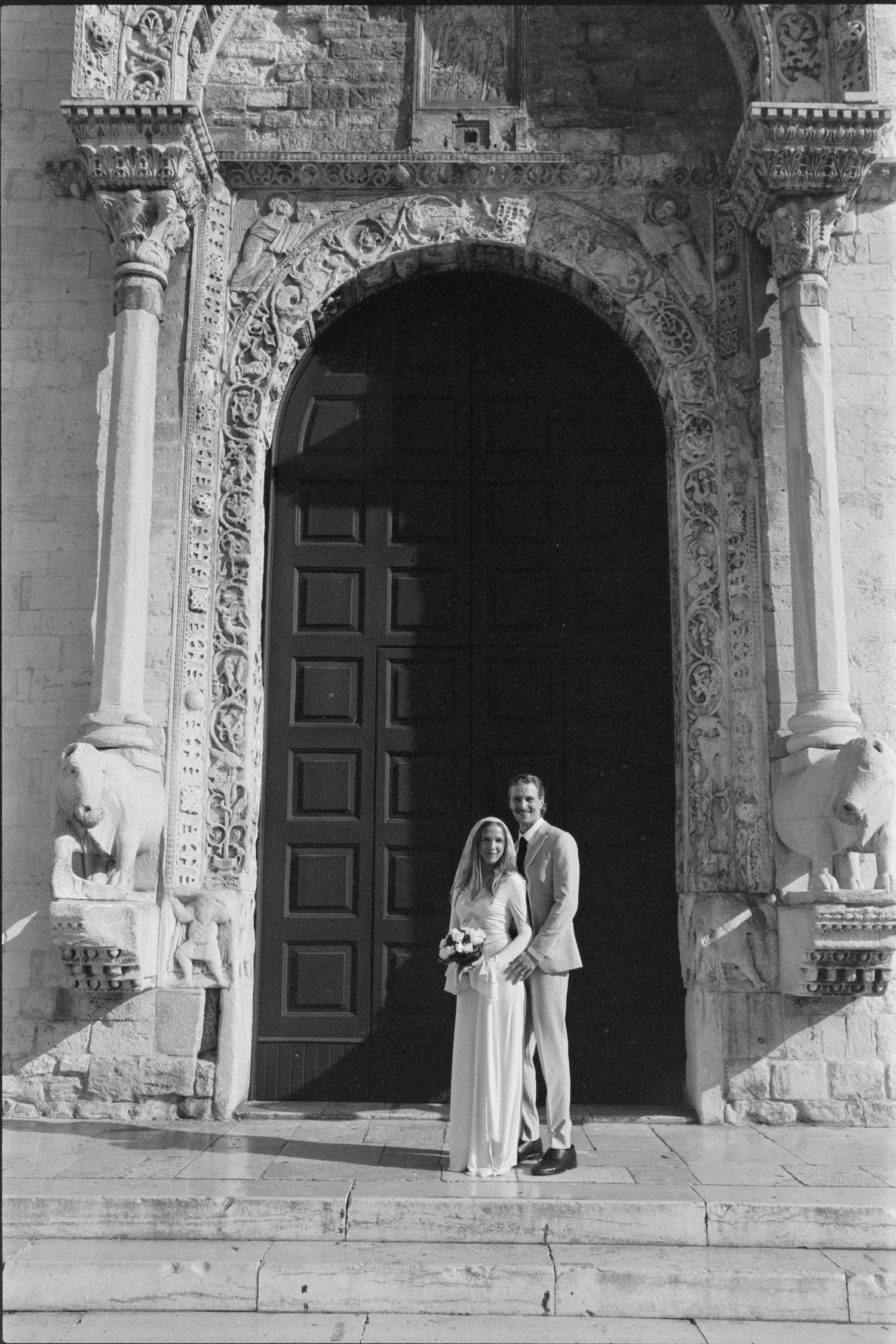 A couple stands in front of an ornate stone archway with decorative columns and a large wooden door at a historic building.