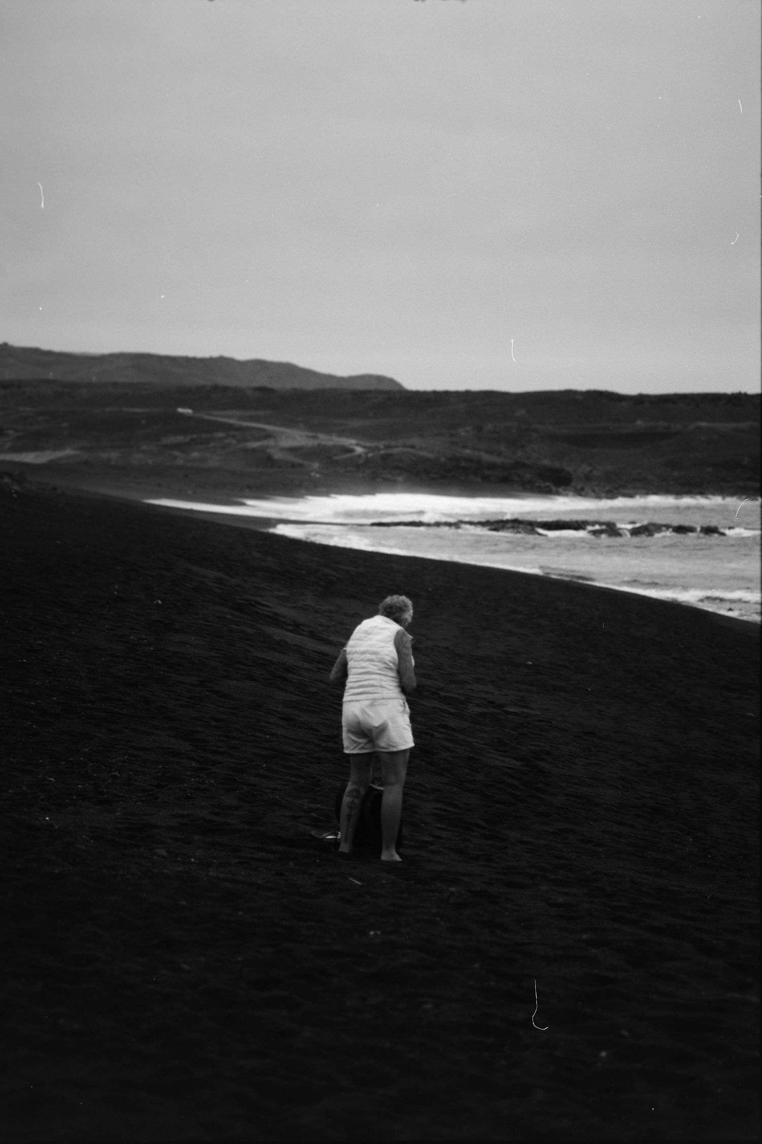 A person in white clothing walks down a steep black volcanic sand beach slope toward distant ocean waves and hills.