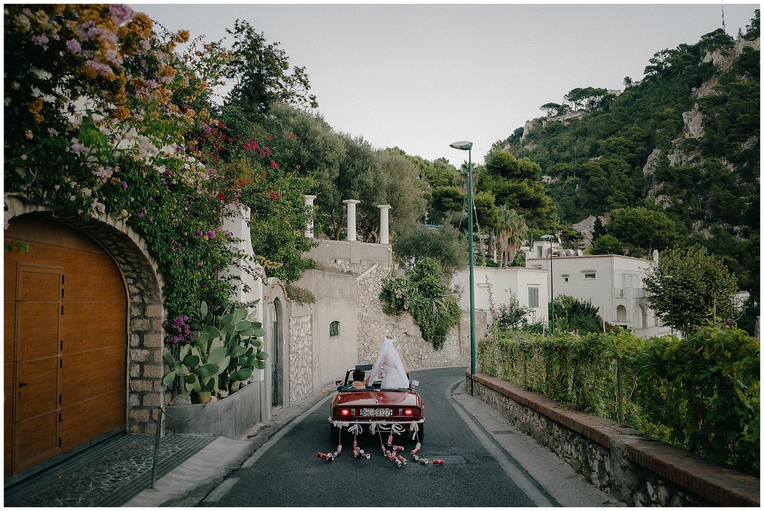 A narrow winding road through a historic Mediterranean village with stone walls and lush greenery on hillside.