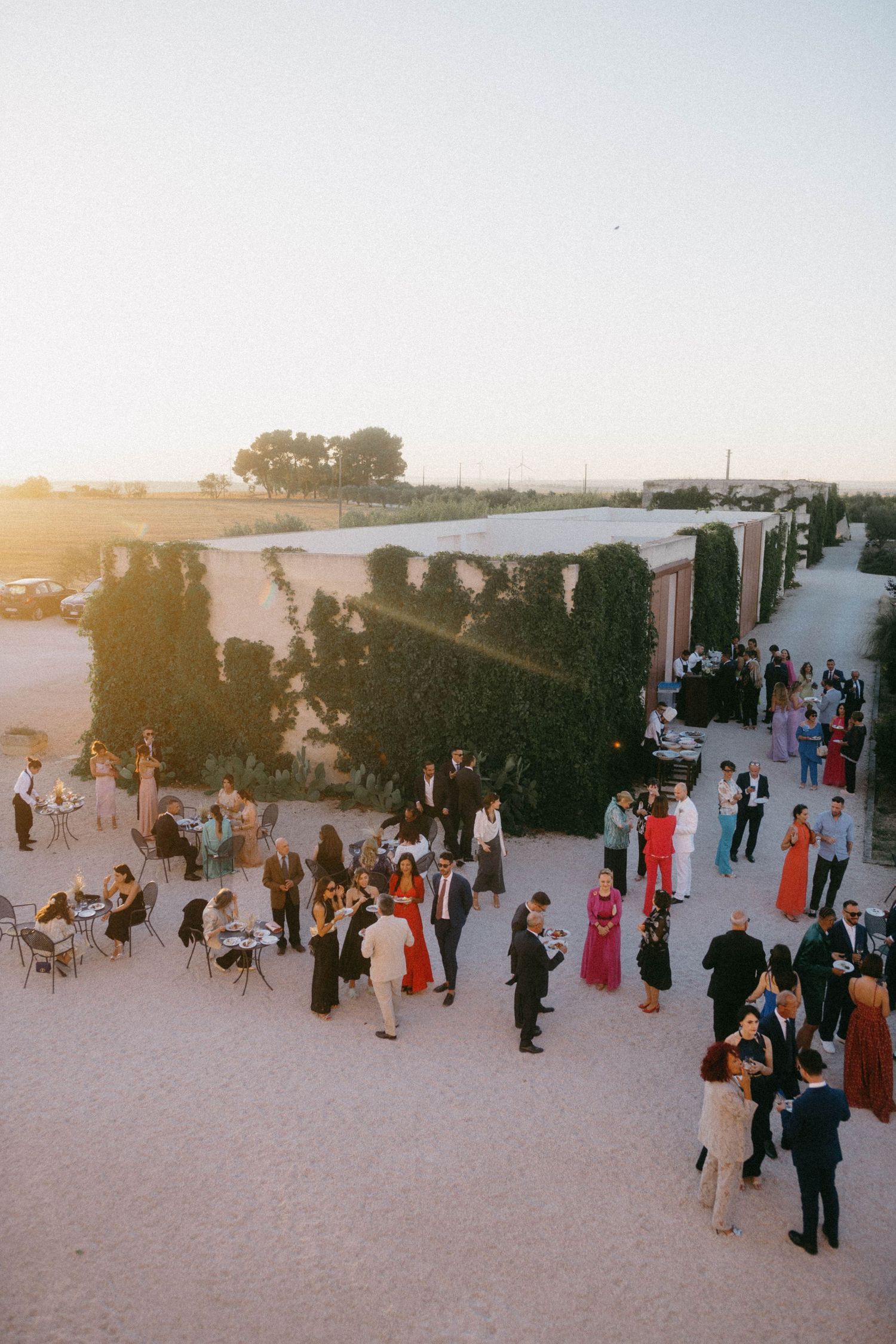 Guests mingling at an outdoor sunset wedding reception on a sandy beach area with trees in the background.