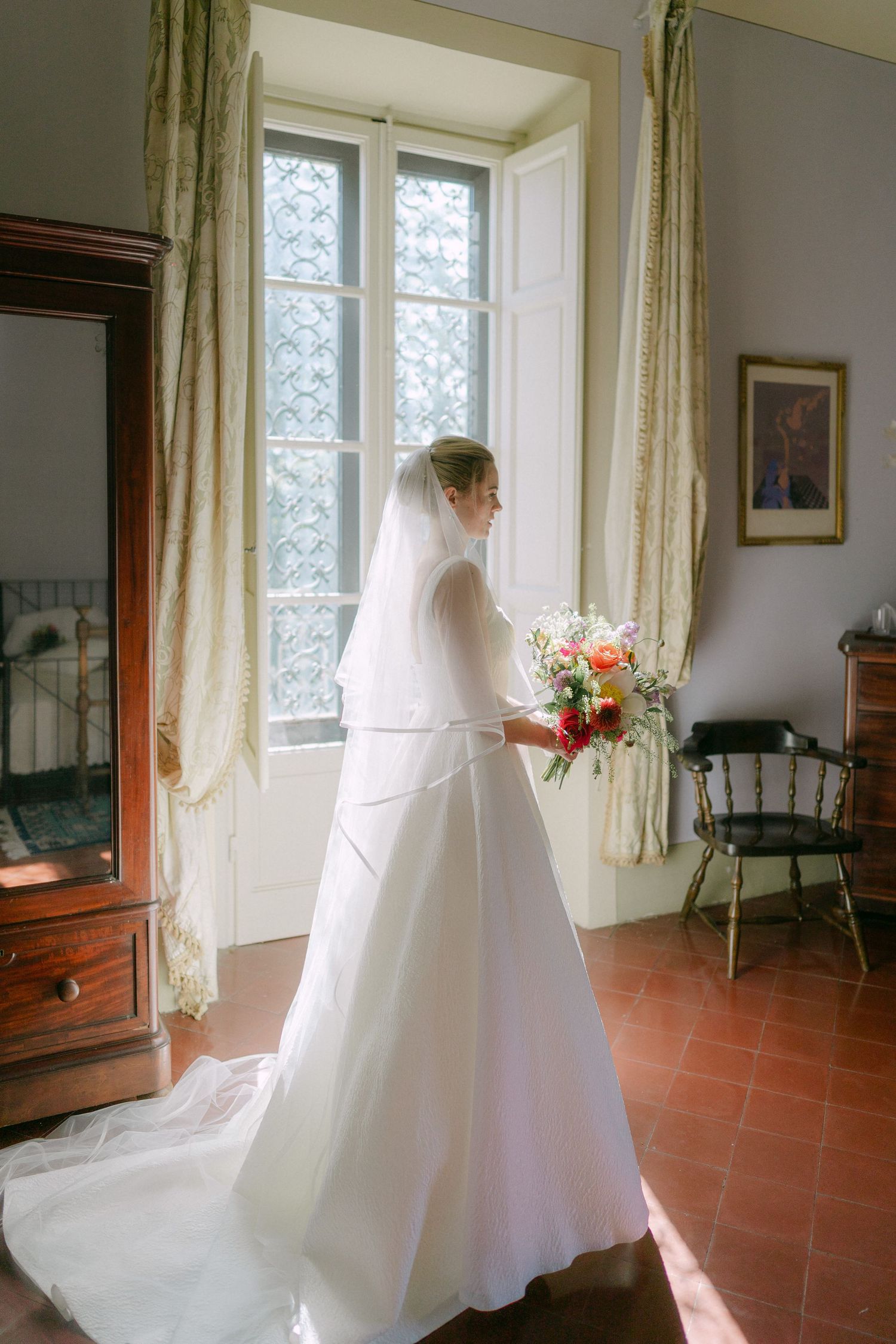 A wedding dress with flowing veil stands elegantly by sunlit windows in a rustic room with terra cotta floors.