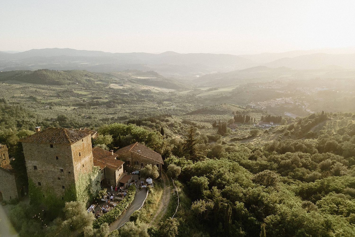 Aerial view of a Tuscan hilltop castle surrounded by lush green trees and rolling valleys in golden sunlight.