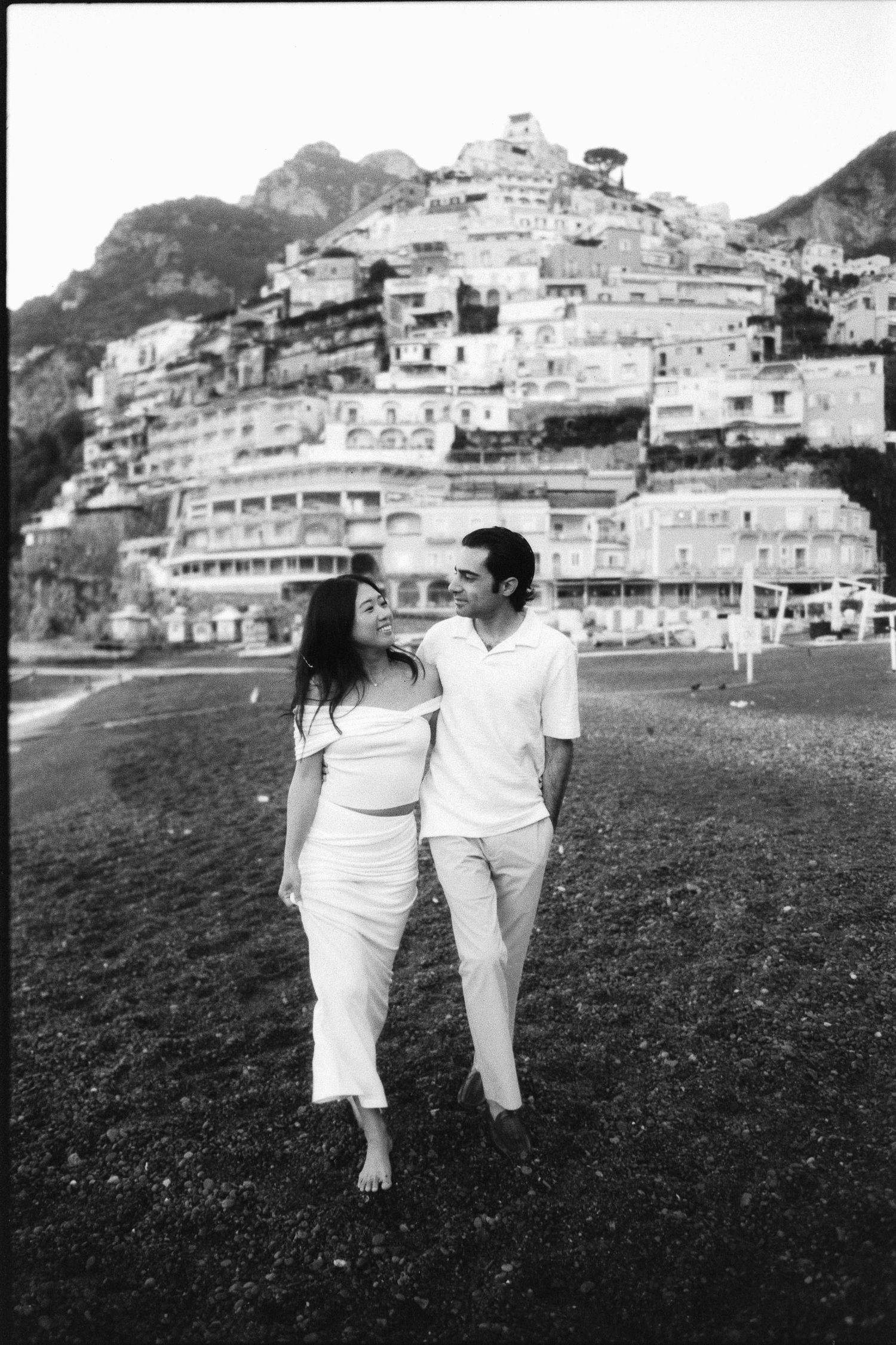 A couple in white outfits walks along the beach by Positano's iconic hillside buildings on the Amalfi Coast in Italy.