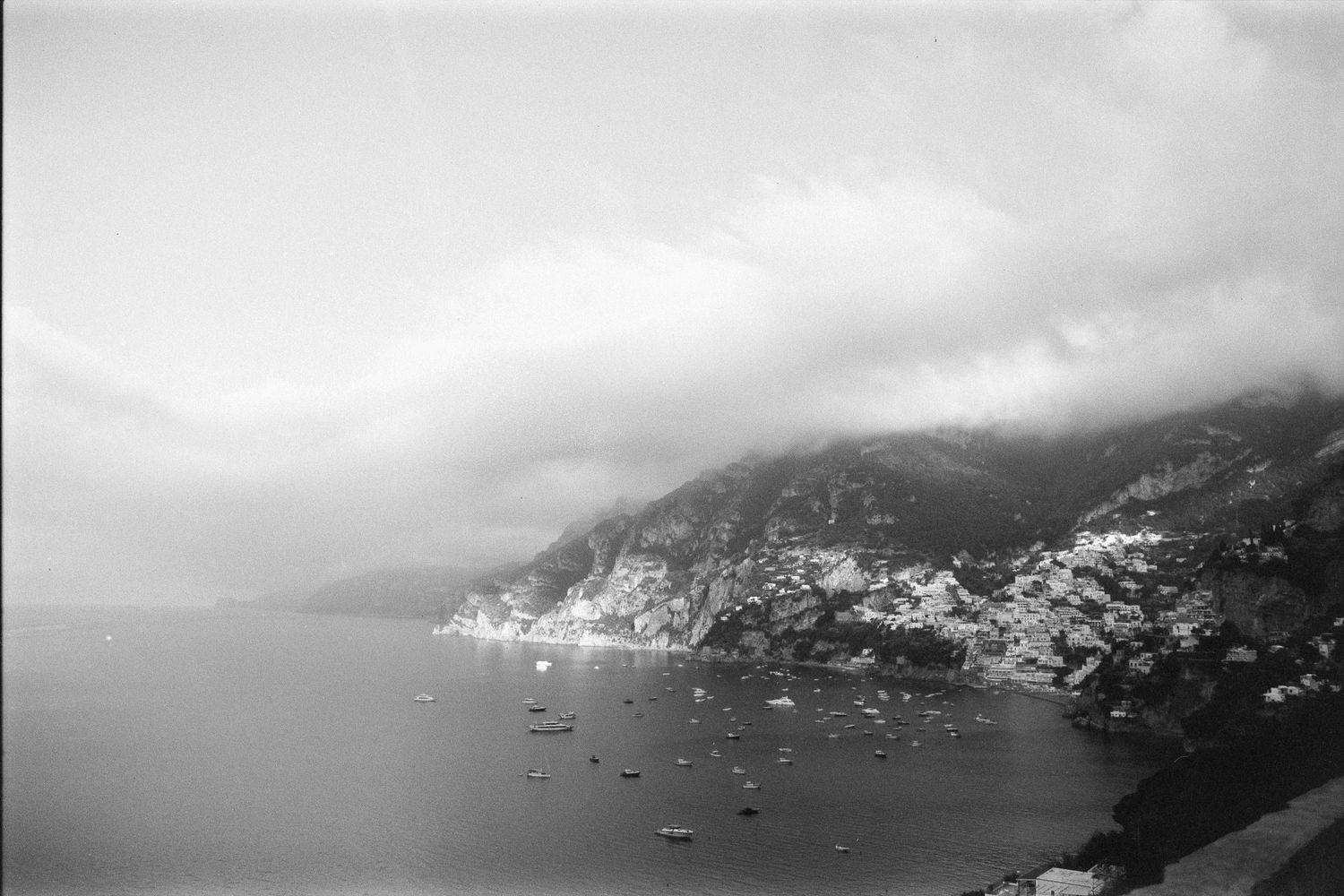 Black and white scenic view of a misty coastal mountainside with boats dotting the Mediterranean waters below.