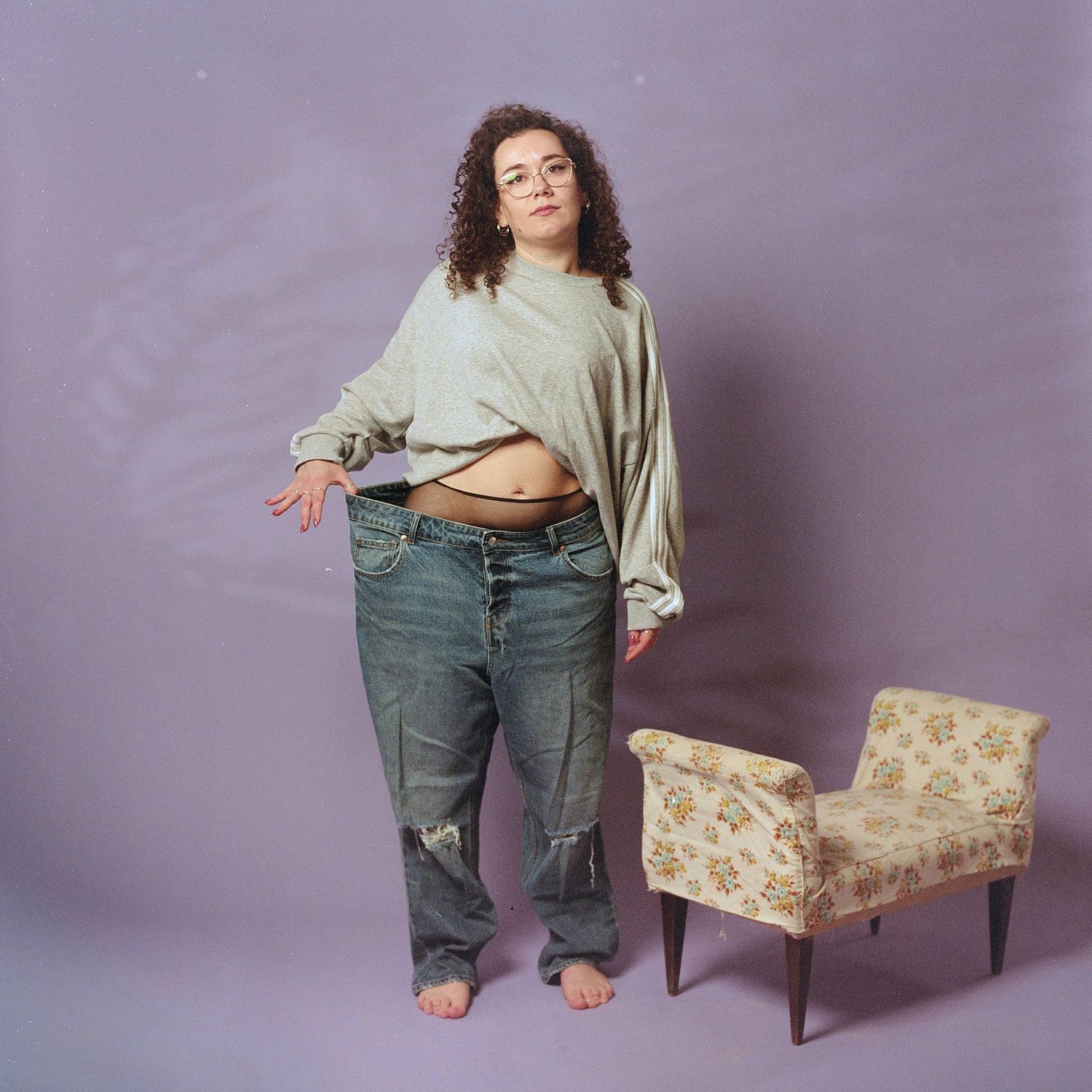 Someone in ripped denim jeans and a cropped beige sweater poses barefoot next to a vintage floral armchair against purple wall.