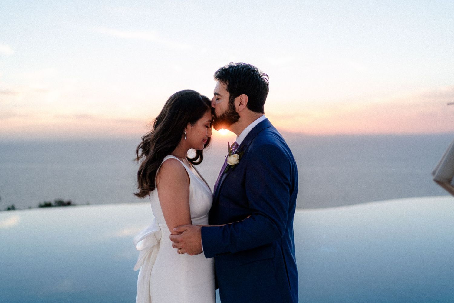 A couple embraces during sunset by an infinity pool overlooking an ocean view.