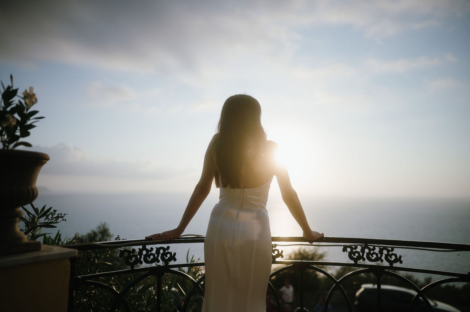Silhouette of woman in white dress leaning on ornate balcony railing at sunset overlooking scenic coastal view.