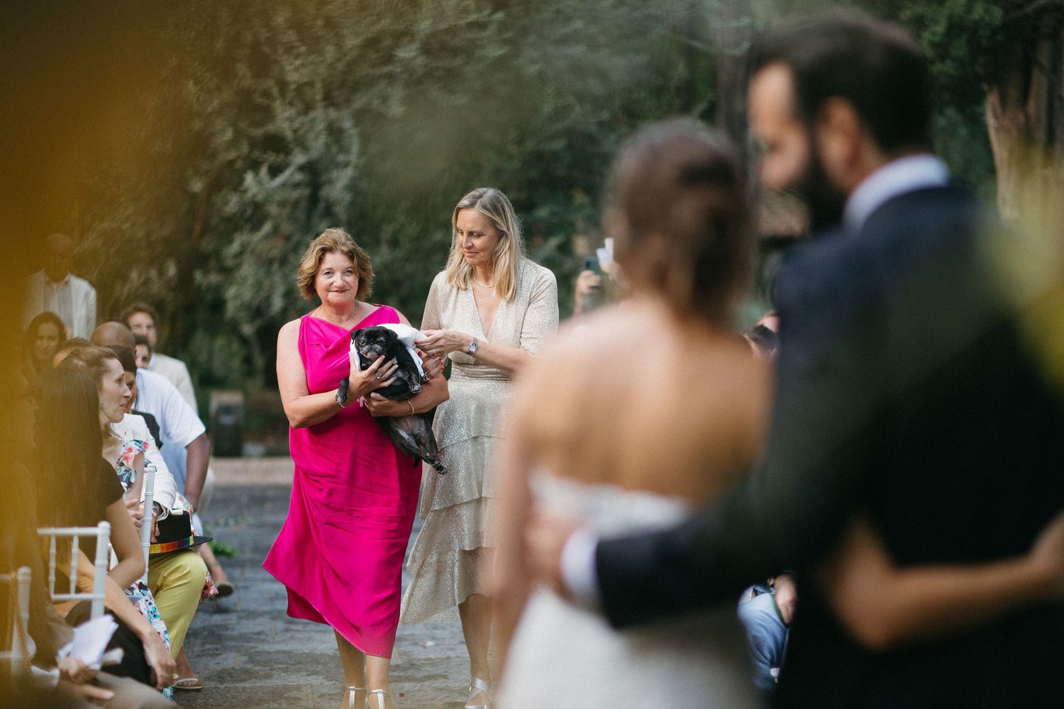 An outdoor wedding ceremony with an officiant in a bright pink dress leading the service in a garden setting.